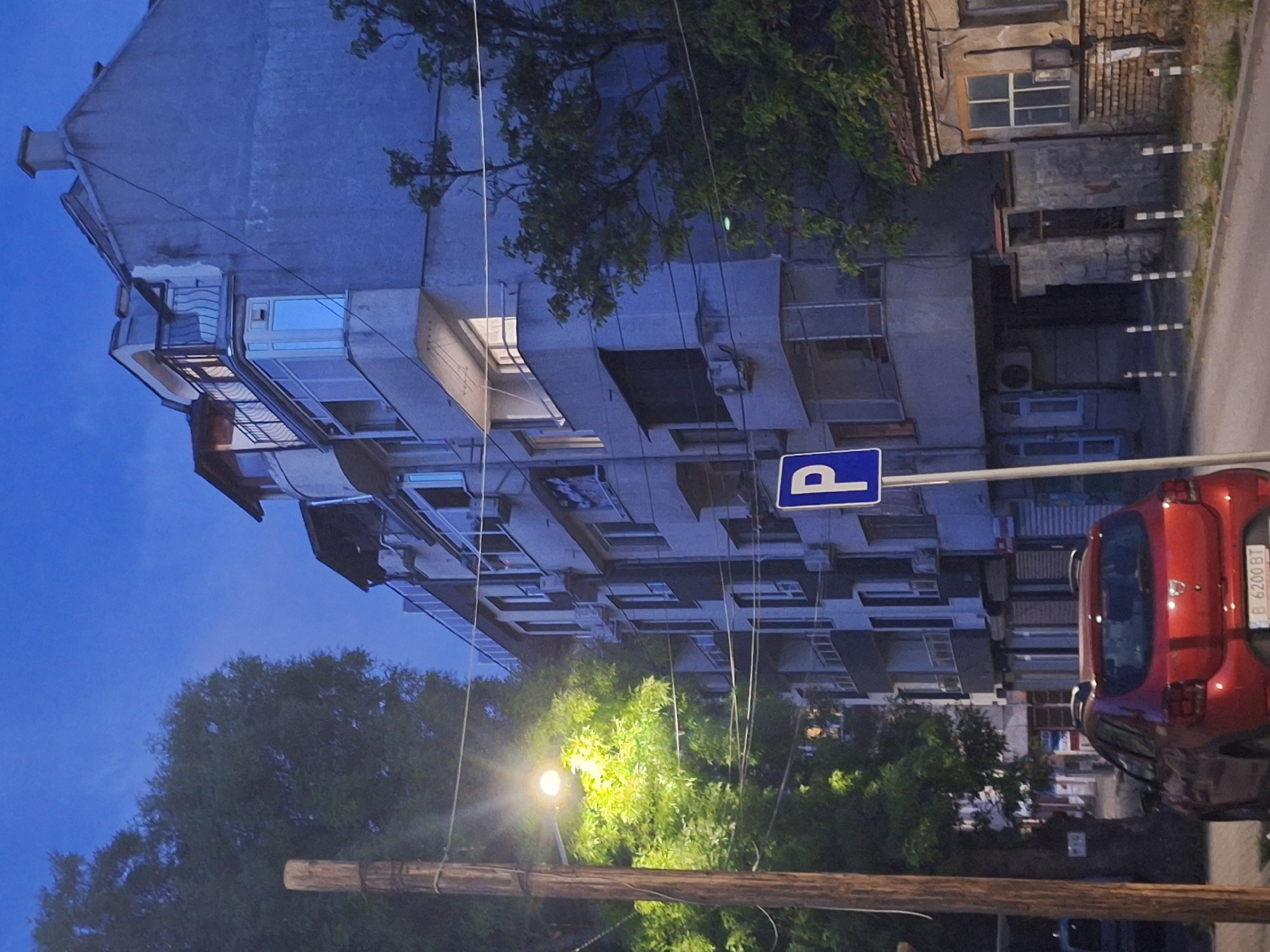 The image depicts a street scene in Varna, Bulgaria, likely during twilight. A multi-story apartment building with a light blue facade dominates the right side of the frame. It features several balconies, some with railings and glass enclosures, and visible air conditioning units. A small, older brick building with a tiled roof and a prominent window is situated to the right of the apartment building.
In the foreground, a red Dacia Sandero hatchback car is parked on the street. A wooden utility pole with a street lamp that illuminates the surrounding foliage stands to the left of the car. A blue parking sign with a white 'P' is visible on a pole near the car. The street is paved, and there are a series of black and white striped bollards along the sidewalk. Trees with green foliage are present, casting shadows. The sky is a deep blue, indicating the late hour. There are no people visible in the image.