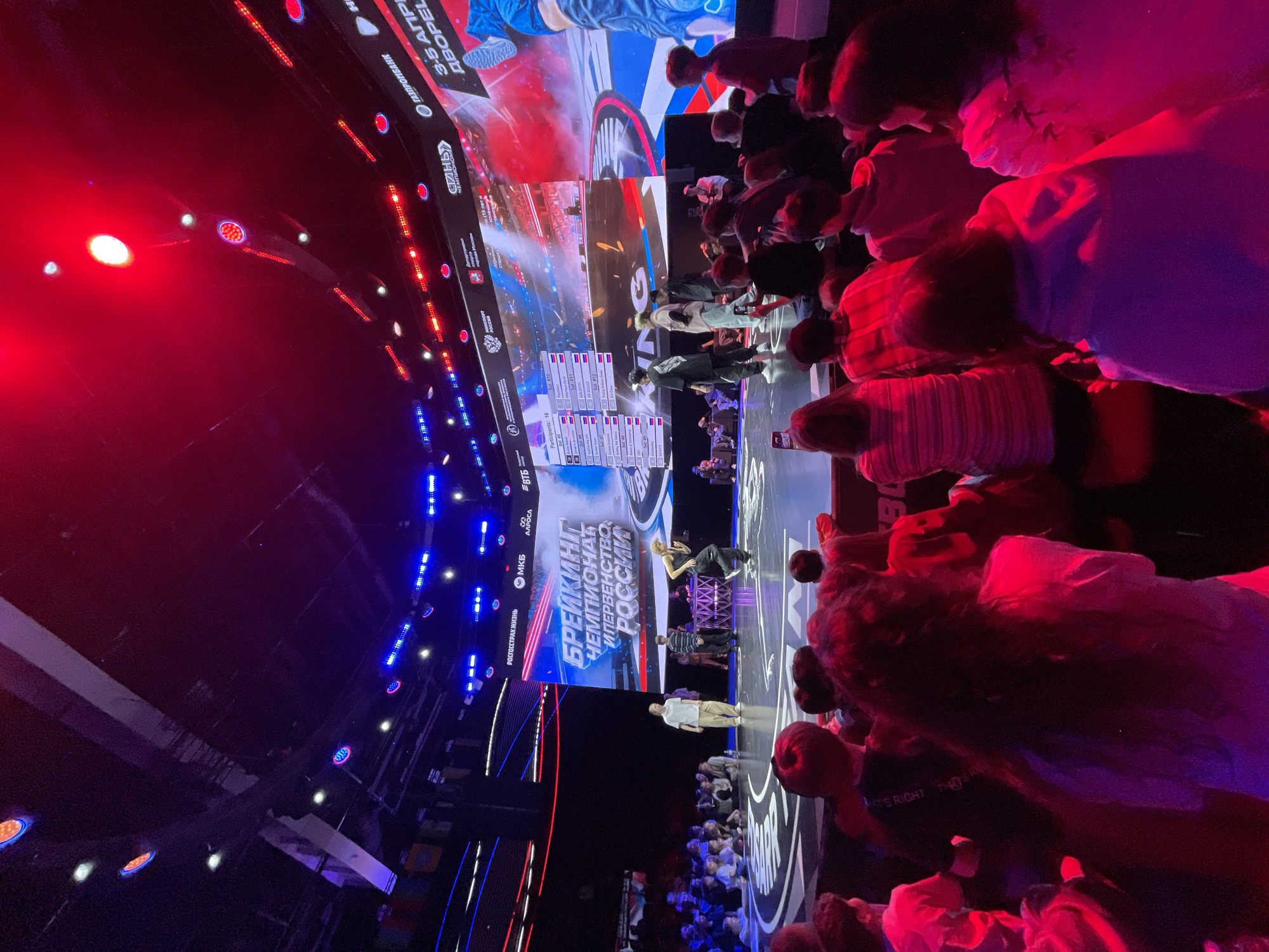 This image shows an indoor event taking place in Moscow, Russia, likely related to a "Breaking Championship." A large stage features illuminated branding and screens. There's a crowd of spectators of all ages and genders gathered in front of the stage, with several people standing. A presenter or performer is on the stage, interacting with participants.