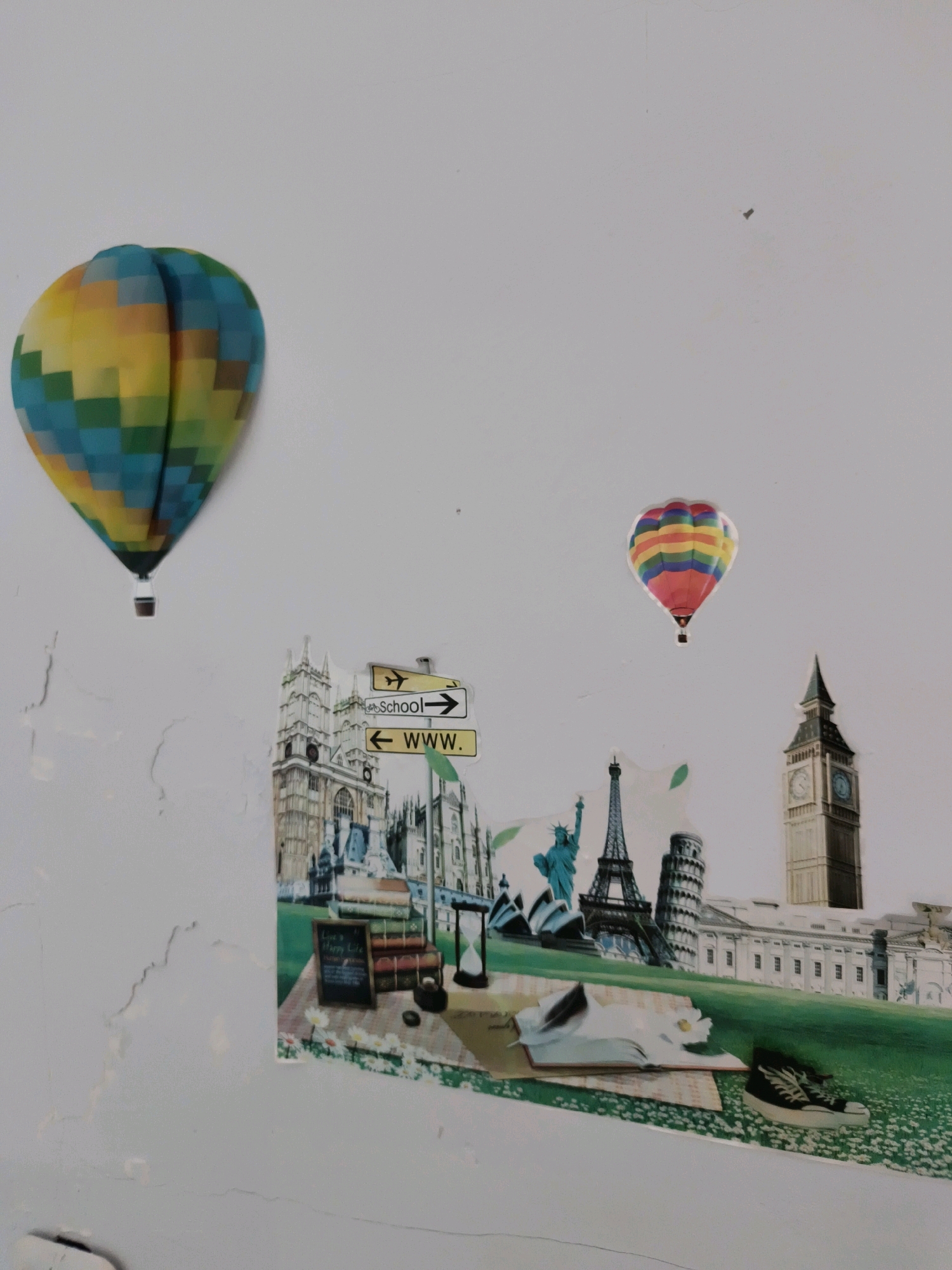 The image shows a collage of travel-themed stickers affixed to a white, textured wall. Two hot air balloons, one larger and multicolor on the left, and a smaller, rainbow-colored one on the right, are depicted. Below them is a scene featuring various international landmarks, including Big Ben, the Eiffel Tower, the Leaning Tower of Pisa, and the Statue of Liberty. There are also signpost stickers indicating "School" with an arrow to the right and "www." with an arrow to the left. In the foreground of the collage, there's an open book, a pair of sneakers, and an hourglass. The wall itself shows signs of wear, with cracks and peeling paint. The location in the city of Tombatu Dua, Indonesia, is not discernible from the image.