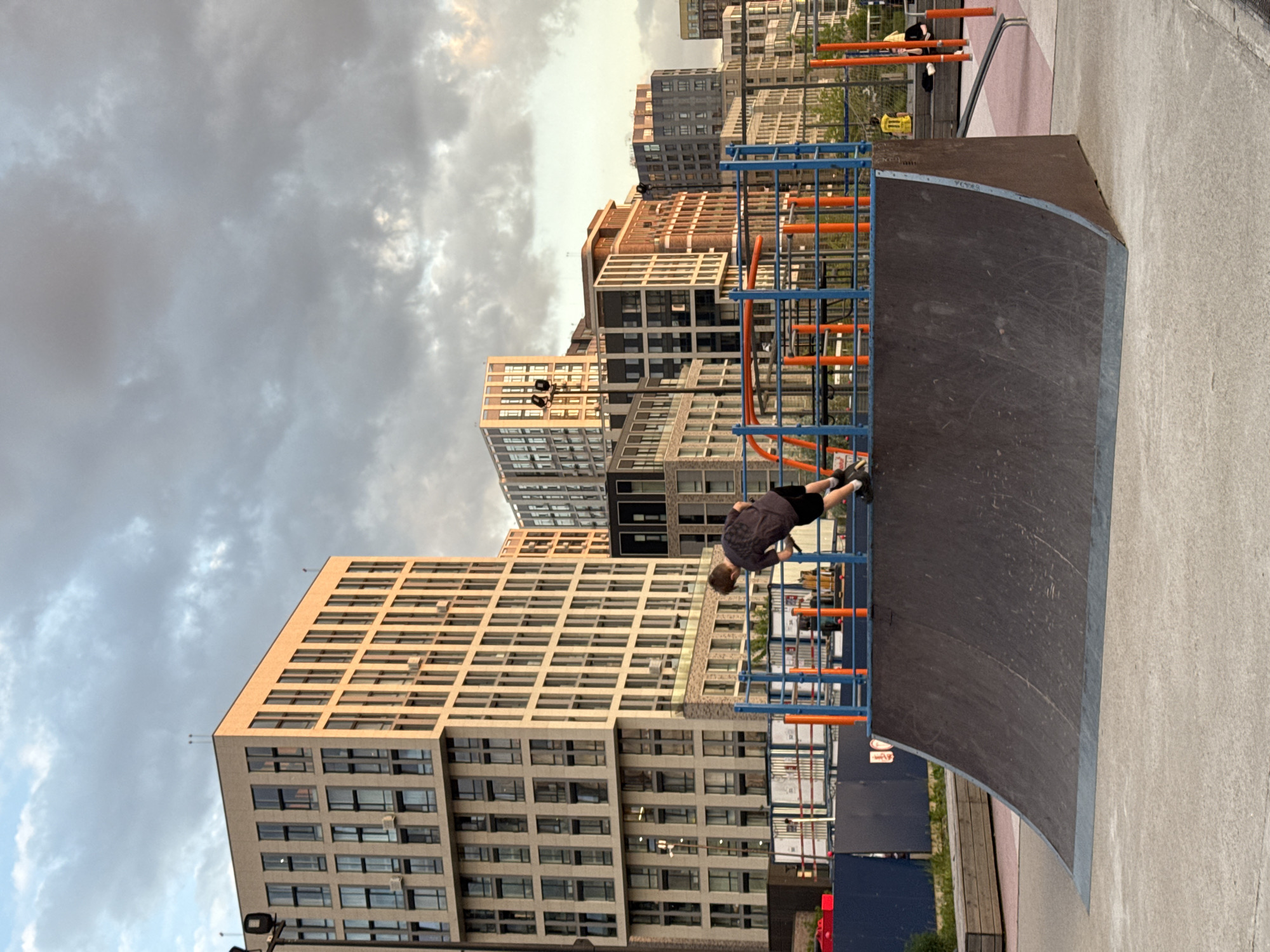 The image captures an urban skate park or recreational area in Moscow, Russia, during what appears to be late afternoon or early evening. The sky is partly cloudy with warm, soft light illuminating the scene.
In the foreground, a large, dark-colored skate ramp or quarter pipe dominates the right side of the frame. Adjacent to it on the left is a blue and orange metal climbing frame or set of parallel bars. A person, appearing to be a young male dressed in a dark short-sleeved shirt and dark shorts, is actively engaged on this structure, leaning forward with their legs raised, performing an exercise or acrobatic move. Further in the background, another individual in a yellow shirt and dark pants is visible near a similar blue and orange structure.
The background is filled with several tall, modern multi-story buildings, characterized by their rectangular windows and varying facades in shades of beige, grey, and brown. These buildings suggest a contemporary residential or commercial urban environment. The ground is paved with concrete. There are no clearly legible texts visible, though a small red and white sign is partially obscured near the base of the skate ramp. The overall atmosphere suggests a lively recreational space within a modern city setting.