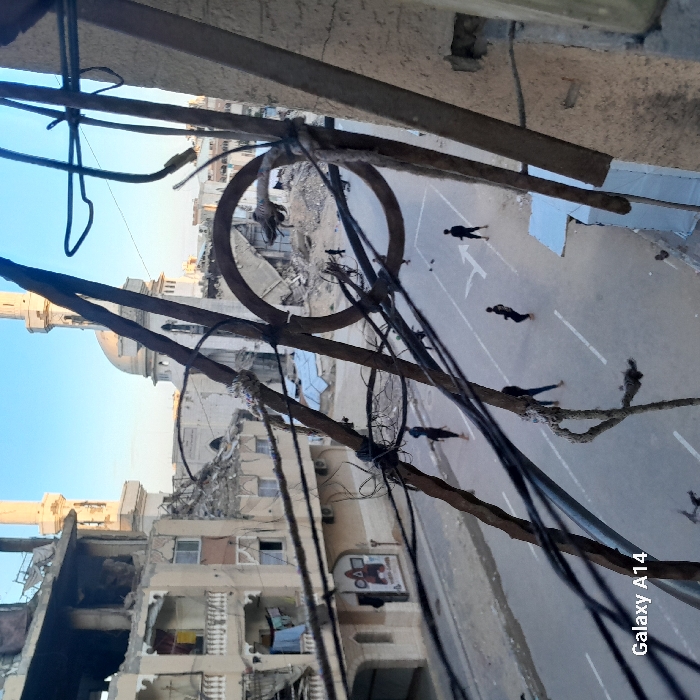 An elevated view captures a street scene in Khan Yunis, Palestinian Territory, characterized by extensive structural damage. In the immediate foreground, rusted metal bars and multiple dark electrical wires, some bound with fabric, obstruct the upper and right portions of the frame.
The midground features a paved street with white lane markings, including solid lines and an arrow indicator. Approximately six to eight individuals, appearing to be young males or children, are dispersed across the street, most dressed in dark clothing; their postures suggest movement or idle standing. To the left, a multi-story building exhibits significant damage, with large sections of its facade and interior exposed, revealing rebar and debris. Laundry hangs from a balcony on this building, and a faded graphic sign is visible on its ground floor.
In the background, a mosque with two minarets and a central dome stands amidst further demolition, with surrounding structures largely reduced to rubble. The mosque itself appears partially damaged or weathered, particularly around its dome and adjacent walls. The sky above is clear and light blue, indicating daylight. The overall environment conveys widespread devastation and ruin across the urban landscape.