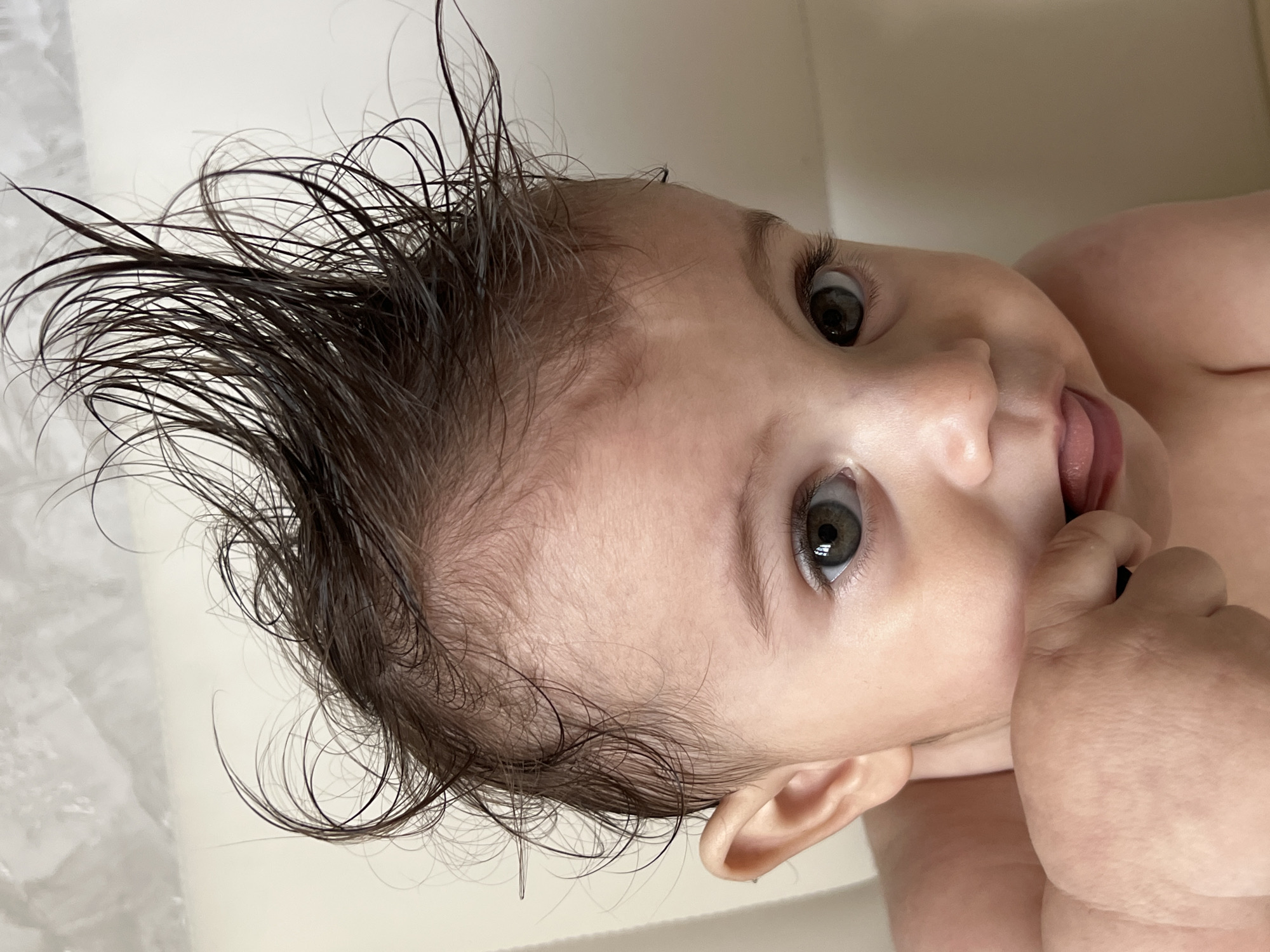 A baby is lying on its back, possibly in a bathtub given the white background. The baby has dark hair that is wet and styled upwards. The baby's eyes are open and focused upward with a curious expression. The baby's right hand is raised to its mouth, with fingers touching its tongue. No location is discernible from the image.