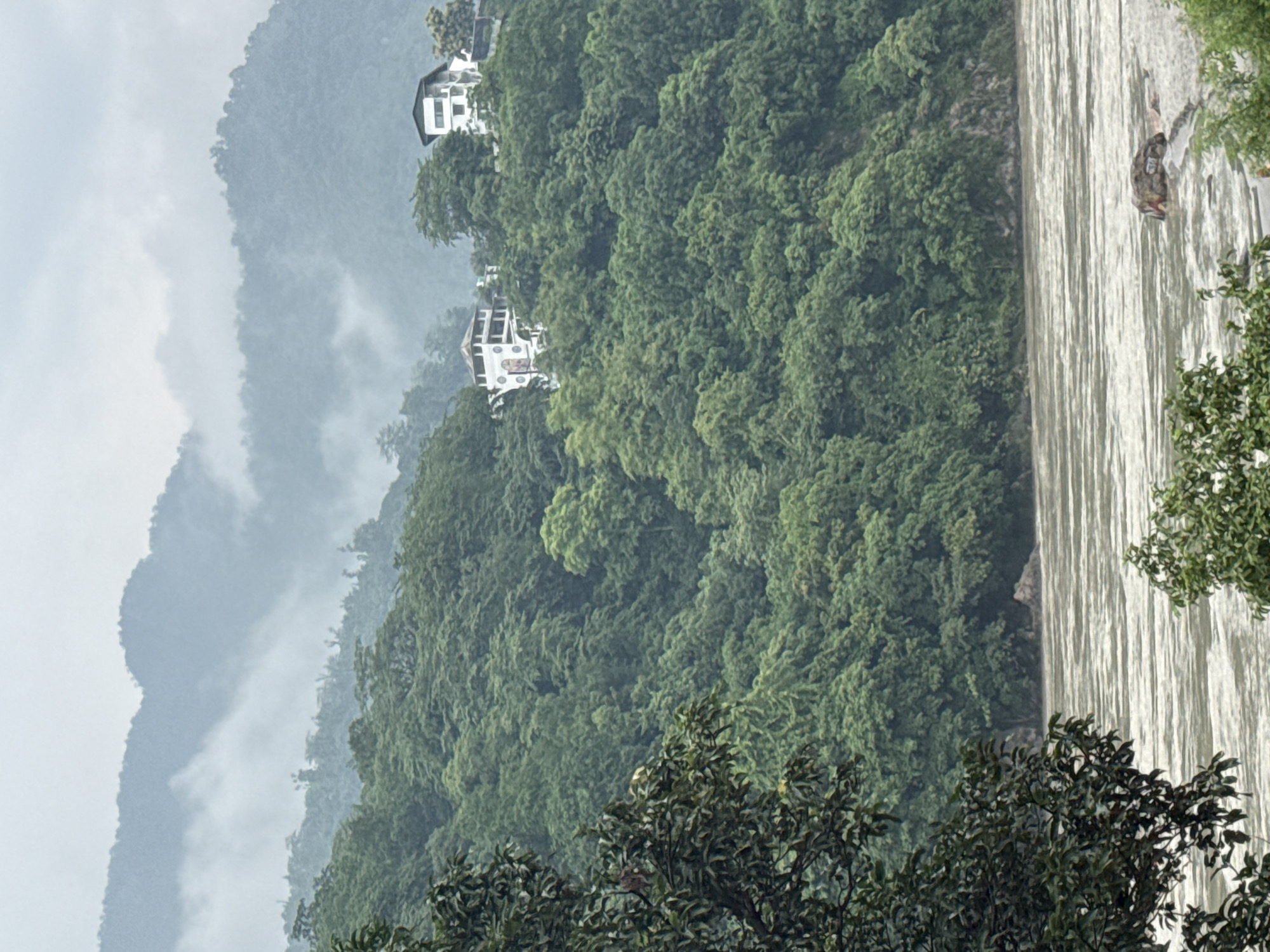 The image shows a wide, turbulent river flowing in the foreground. On the right bank of the river, a large rock formation is visible in the water. The river is surrounded by dense green foliage, suggesting a lush, natural environment. In the background, a steep, forested hillside rises up, with two white multi-story buildings nestled amongst the trees near the top of the hill. The sky is overcast and hazy, with faint outlines of distant mountains visible through the mist. The scene is likely located in or near the city of Rishikesh, India, a region known for its hilly terrain and the Ganges River.