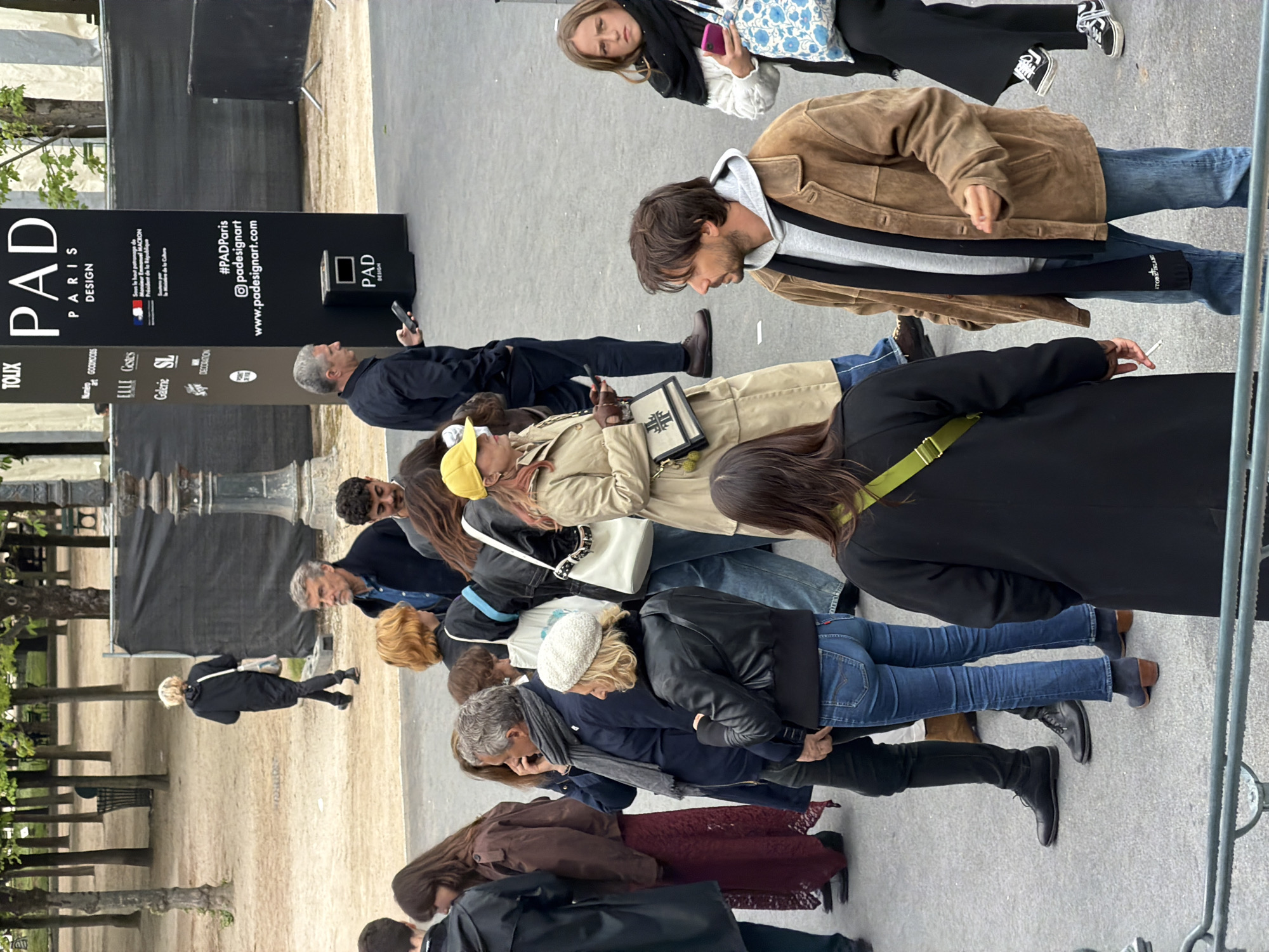 A slightly tilted, high-angle, medium shot captures a diverse group of people, some gazing at or interacting with the PAD Paris Design exhibition, which is signified by a large black sign displaying the event's name, sponsors, and website. The crowd is on a grey stone walkway adjacent to a dirt path scattered with trees and metal benches in the city of Paris, France.
The people exhibit a range of ages and wear casual attire, including jeans, jackets, and hats. Several individuals are holding mobile phones, and at least one person is holding a book-shaped object. Their expressions suggest attentiveness or focused interest in the exhibit.
The black exhibition signs provide event information, listing #PADParis and the event's website. The sign also mentions that the event is under the patronage of Emmanuel Macron.