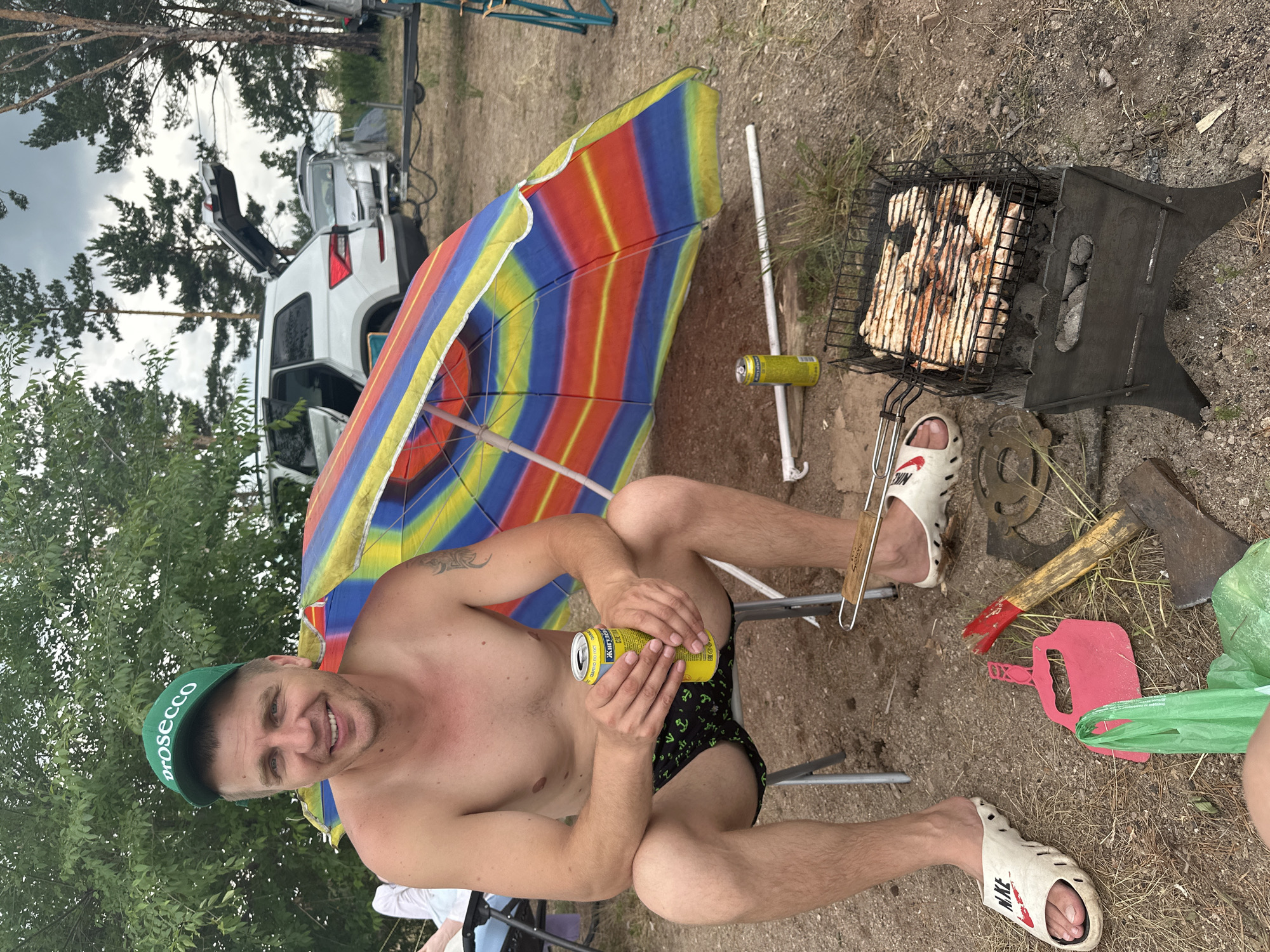 In the city of Zhemchuzhny, Russia, a shirtless man wearing a green cap that says "Prosecco" and black swim trunks with an anchor pattern sits in a folding chair next to a colorful beach umbrella. He is smiling and holding a yellow can of what appears to be a beverage. In front of him, a small grill with chicken pieces cooking over coals is visible. Various items are scattered around the ground, including a red dustpan, an axe, a green plastic bag, and two cans. In the background, several vehicles, including a white SUV with its trunk open and a silver car, are parked in a wooded area. The scene suggests an outdoor gathering or picnic.
