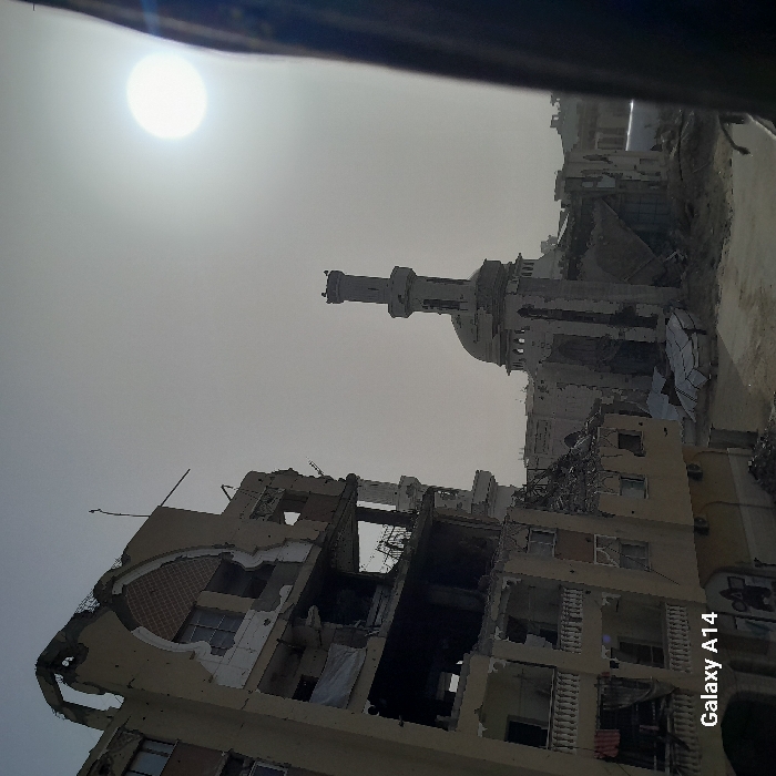 War-torn buildings and a mosque minaret stand against a hazy sky with a bright sun. Buildings show significant structural damage. The mosque appears damaged, with the minaret still standing. Rubble and debris are scattered around. The scene is filled with destruction and devastation.