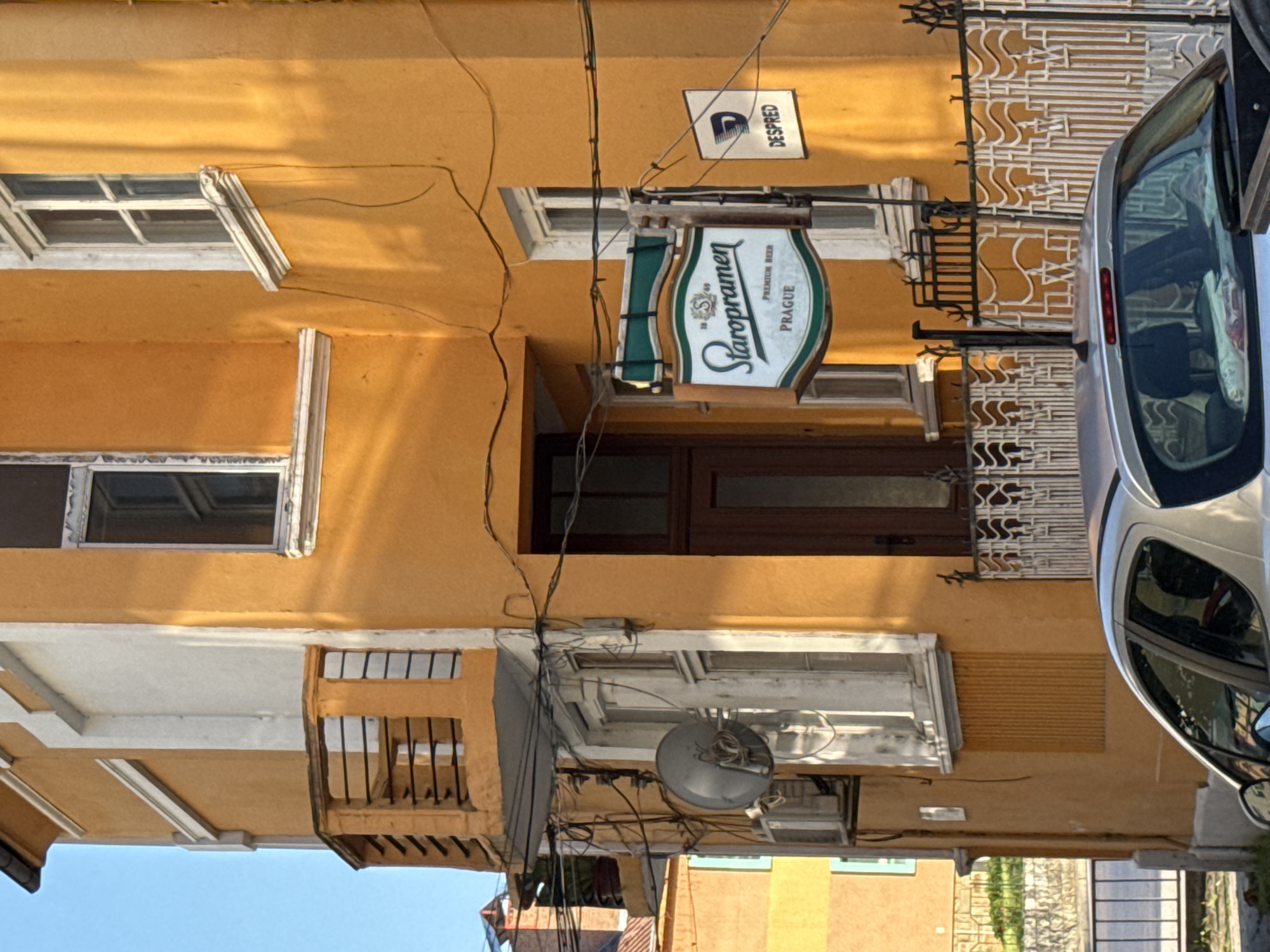 The image shows a building in Lovech, Bulgaria, with a "Staropramen" beer sign hanging above a doorway. The building is yellow with white trim around the windows and a small balcony. A satellite dish is mounted on the building's exterior. A silver car is parked partially in front of the building, obscuring some of the facade. Power lines are visible running across the building's exterior. A sign labeled "DESPRED" is also present. The sky is visible in the lower left corner of the image.