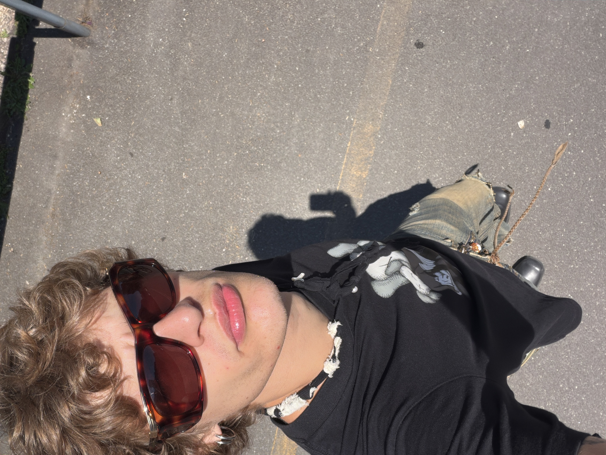 A person is lying on an asphalt surface outdoors, possibly a road or pavement. The asphalt has a yellow painted line on it. The person wears brown tortoiseshell sunglasses, a black t-shirt with white cartoon graphics, and blue jeans. A braided whip is visible near their leg. A metal bar is in the background to the upper left. Based on the urban asphalt environment and the style of the person, the location is likely within a city, possibly Milan, Italy.