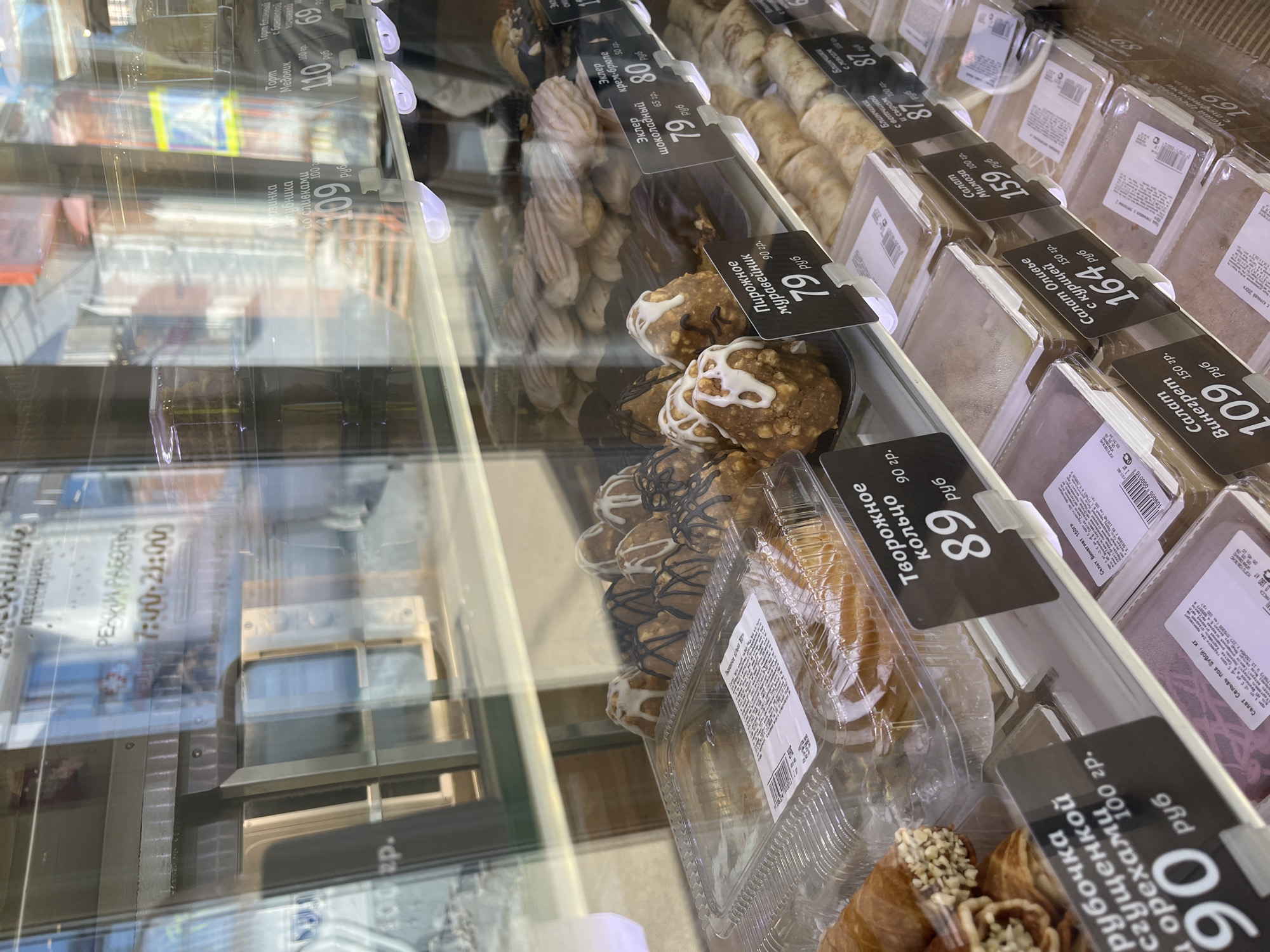A refrigerated display case holds various pastries and prepared salads for sale in a shop in Izhevsk, Russia. The items are individually packaged in clear plastic containers, with price tags in front. The pastries include cream puffs, eclairs, and cheese rings. The salads, in boxes, are vinaigrette and mimosa salad. The prices are displayed in Russian rubles. The image is taken through the glass of the display case, reflecting the building across the street and the shop's hours of operation, 7:00 AM to 9:00 PM.