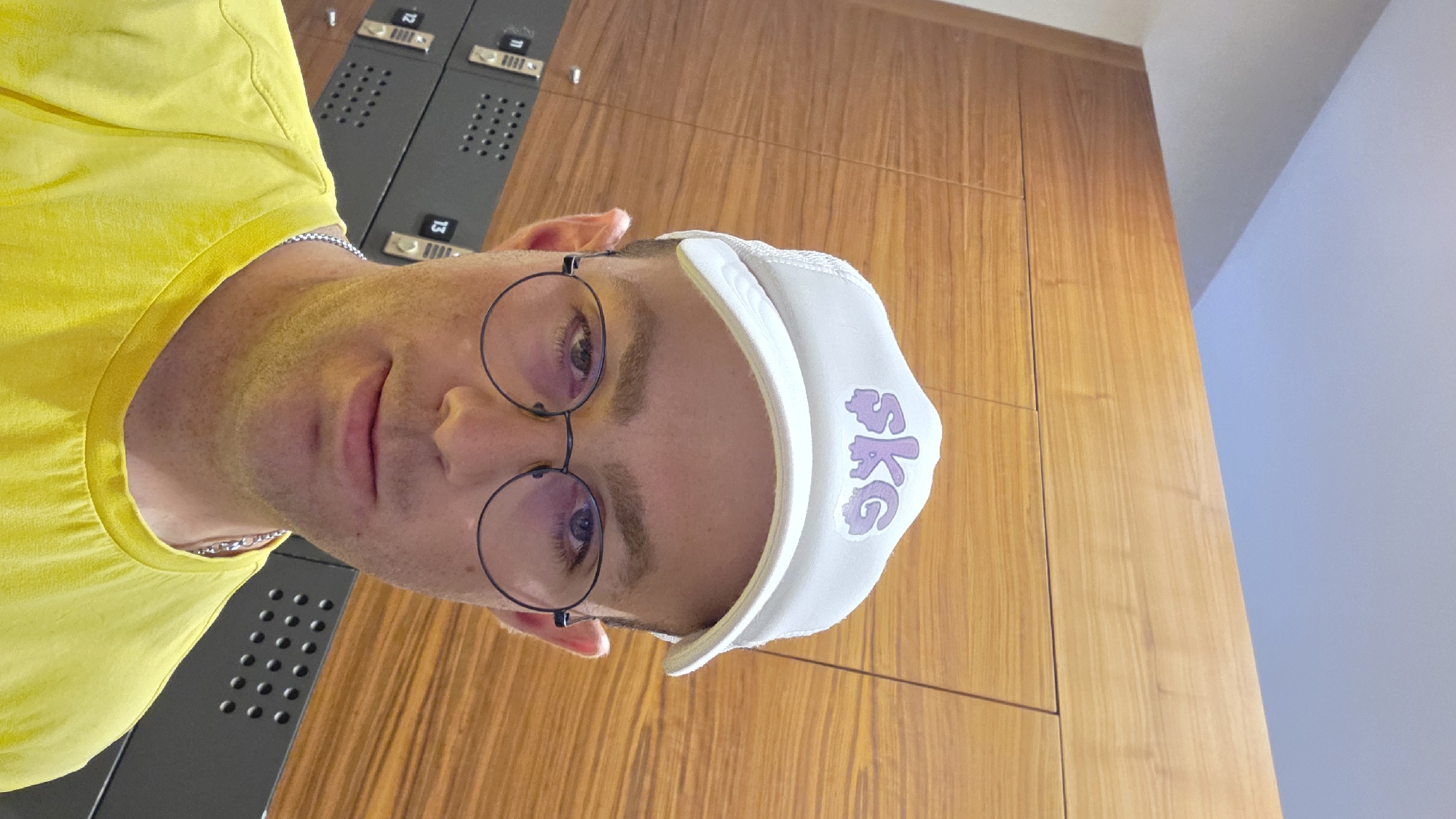 A close-up selfie of a man wearing a white trucker hat with a purple "skg" logo and round, black-rimmed glasses. He is also wearing a bright yellow t-shirt and a silver chain. He appears to be in a locker room with wooden paneling and grey lockers in the background. The lockers have visible numbers, including "11", "12", and "13". The lighting is bright, suggesting the image was taken indoors. The location is identified as Bania, Bulgaria.