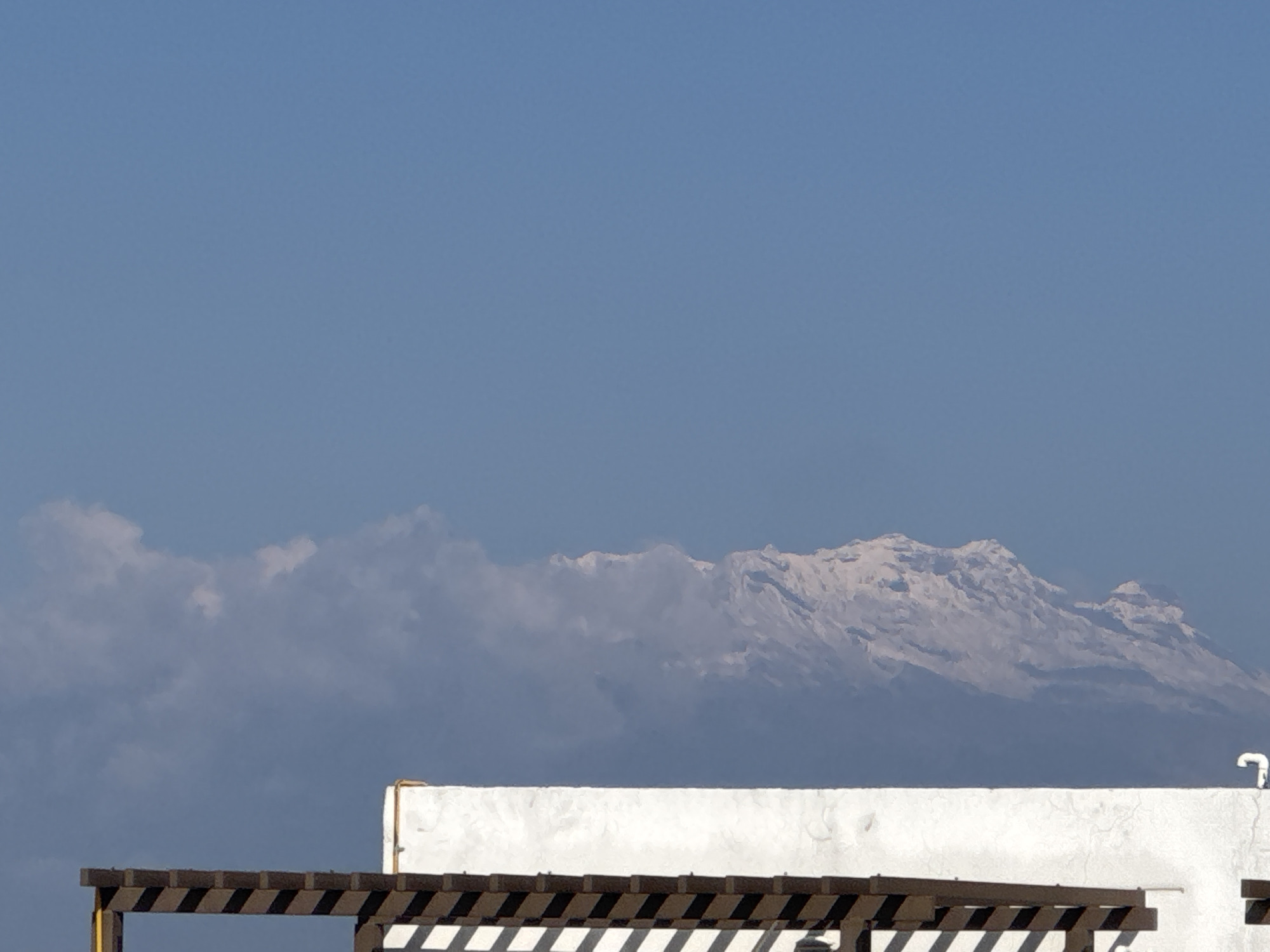 The image shows a daytime view of a snow-capped mountain range under a clear blue sky, likely the Iztaccihuatl stratovolcano, as seen from San Francisco Ocotlán, Mexico. A white wall and a wooden pergola are in the foreground. The scene appears static and unoccupied. A white curved pipe protrudes from the white wall.