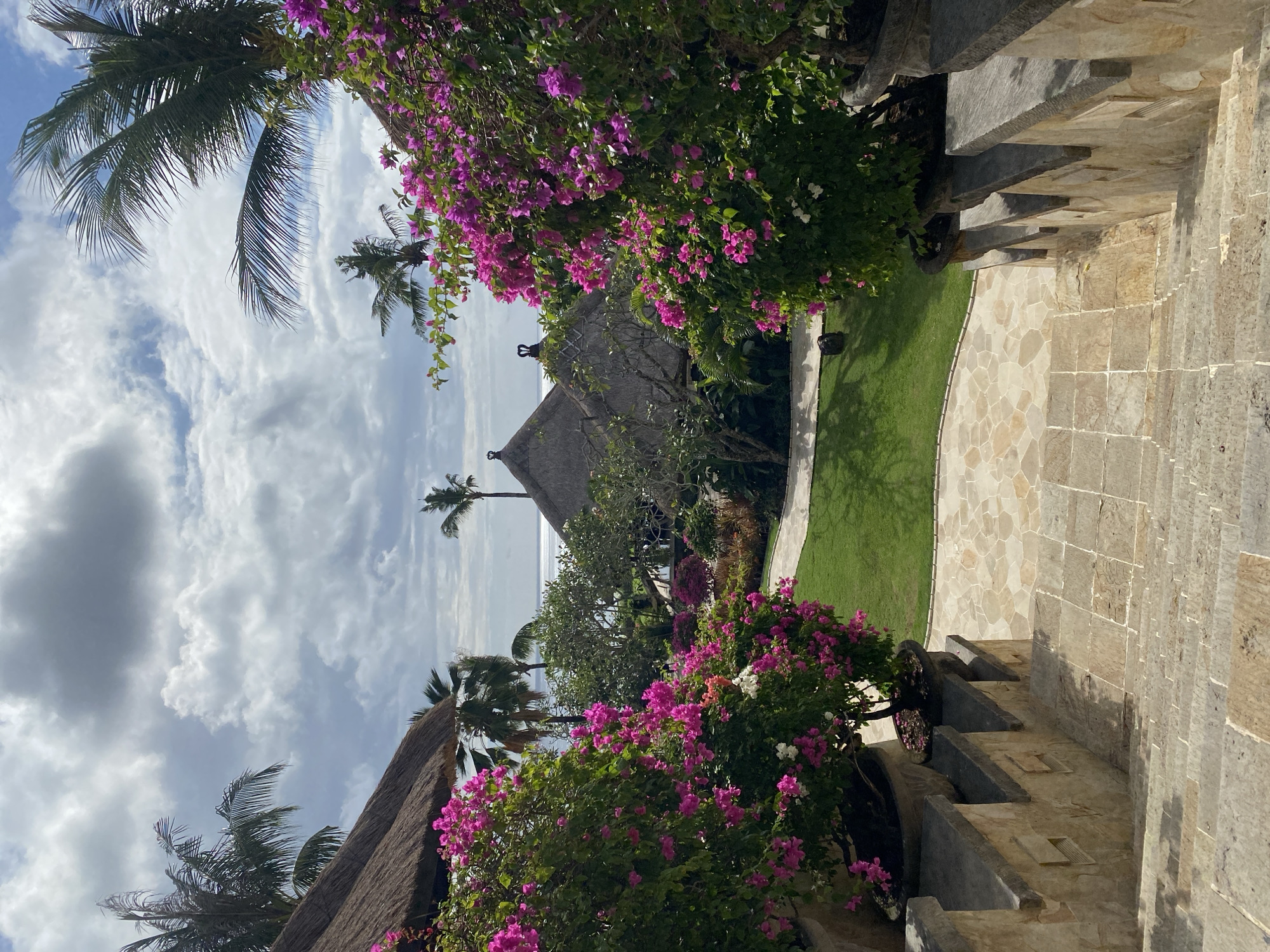 This media file depicts a scenic view of a tropical resort or private residence in Kuta Selatan, Indonesia. The scene is characterized by lush vegetation, including palm trees and vibrant pink bougainvillea flowers in full bloom, which frame the view. In the background, thatched-roof structures, possibly bungalows or villas, are visible, hinting at accommodation. Beyond the structures, a calm ocean stretches towards the horizon under a sky filled with dynamic clouds.
The composition suggests a pathway or stairway made of stone leading down into the property, bordered by manicured green lawns. The stonework appears aged, adding to the rustic charm of the location. The bright sunlight and fluffy clouds indicate it is daytime, likely mid-morning or afternoon, with pleasant tropical weather.
No people are visible in the frame, emphasizing the tranquility and serene atmosphere of the location. The overall impression is one of tropical paradise, perfect for relaxation and enjoying the natural beauty of the surroundings. The image evokes feelings of peace and escape. There is no visible text in the media file.