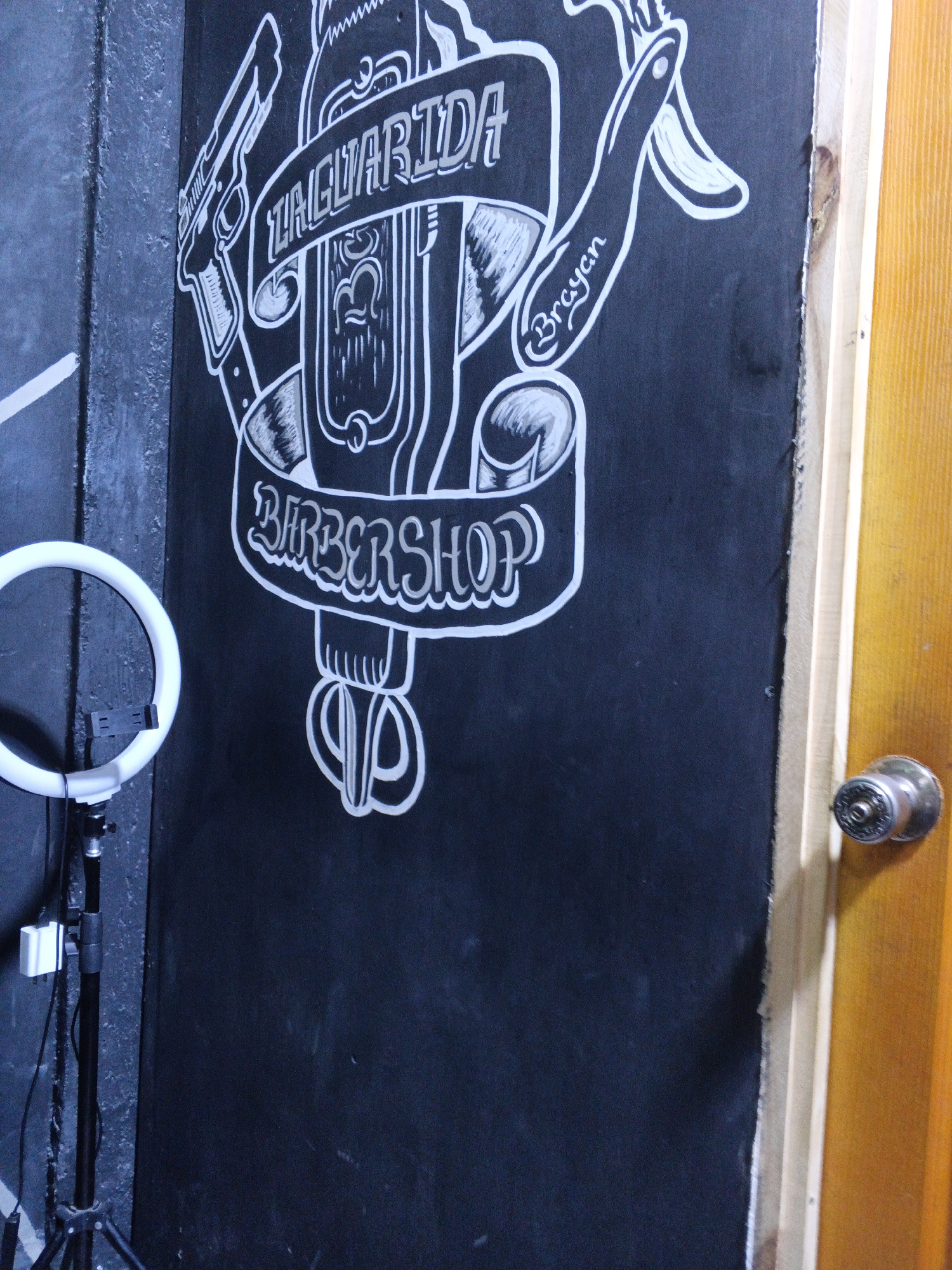 The image depicts a black board with white graffiti-style artwork that advertises a barbershop called "La Guarita Barbershop," with the name "Brayan" also written. The artwork features a gun, a straight razor, and barber tools. To the left of the board is a ring light on a stand. To the right is a door with a silver doorknob. The image was taken in the city of Altagracia, Venezuela.
