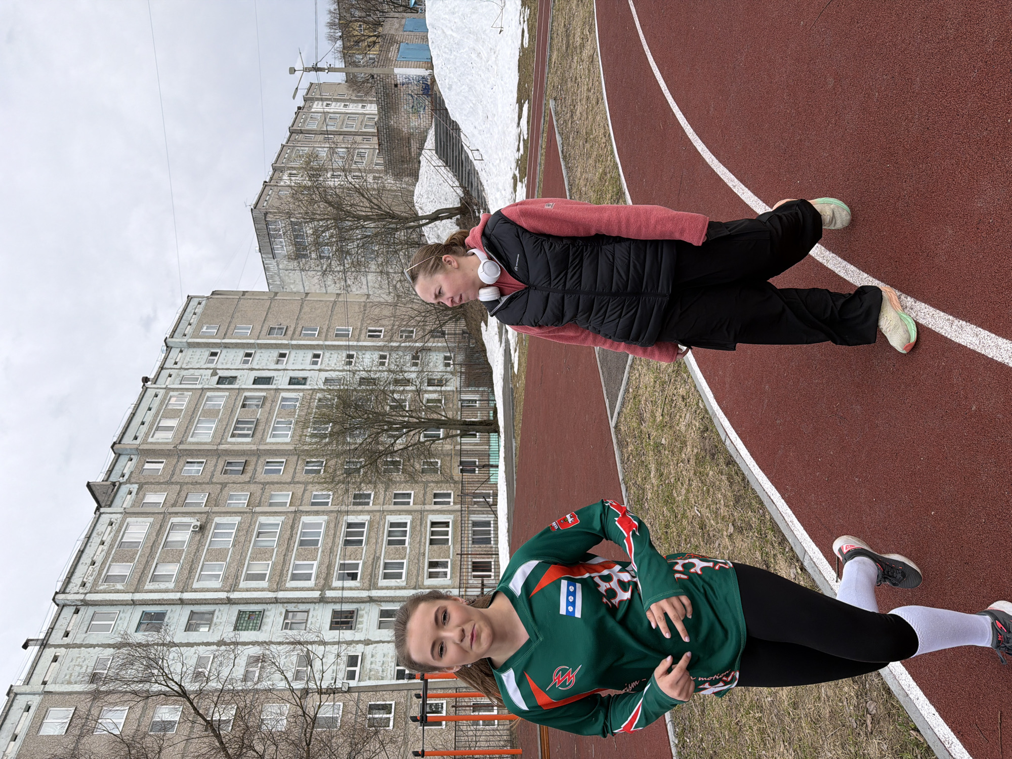 Two young women are depicted on an outdoor running track in an urban setting, likely in Berezniki, Russia, during an overcast day in late winter or early spring. Patches of snow are visible on the ground, and trees in the background are bare, suggesting the season.
In the foreground, a woman with dark hair looks towards the camera with a slight smile. She is wearing a dark green long-sleeved athletic shirt adorned with white and orange accents, including a stylized "W" or "M" logo on the chest, a flag-like emblem with three blue squares on a white background, and animal-print patterns on the shoulders. She pairs this with black leggings, white mid-calf socks, and dark athletic shoes. Her hands are held in a relaxed, casual pose.
Further back on the track, another woman with her hair pulled back wears a pink or light red hooded top under a black puffer vest, black pants, and light-colored athletic shoes with distinctive soles. She has white over-ear headphones resting around her neck and is looking downwards and to her right.
The running track is red with clear white lane markings. Beyond the track, the ground consists of dry grass and dirt, interspersed with melting snow. The background features several large, multi-story concrete apartment buildings, characteristic of Soviet-era architecture, reinforcing the geographical context. Bare trees stand between the track and the buildings. The overall scene suggests a casual outing or break on the track rather than active exercise.