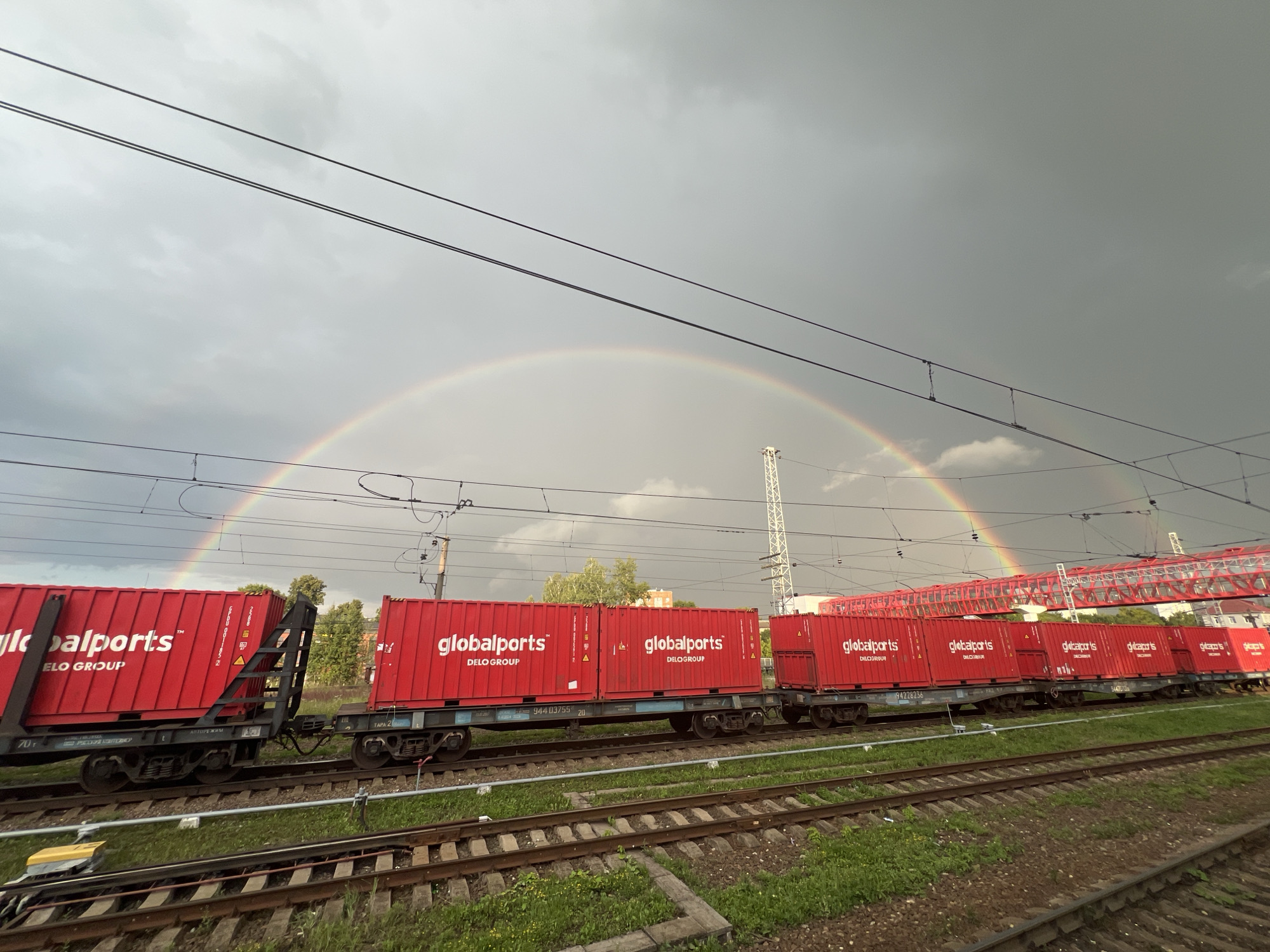 A train carrying red shipping containers with the "globalports DELOGROUP" logo is on tracks under a stormy sky. A rainbow arcs across the sky, partially obscured by clouds and overhead power lines. In the background, buildings and a red pedestrian bridge are visible. The location is Serpukhov, Russia. The scene appears to be a freight yard or industrial area. No people are visible in the image.