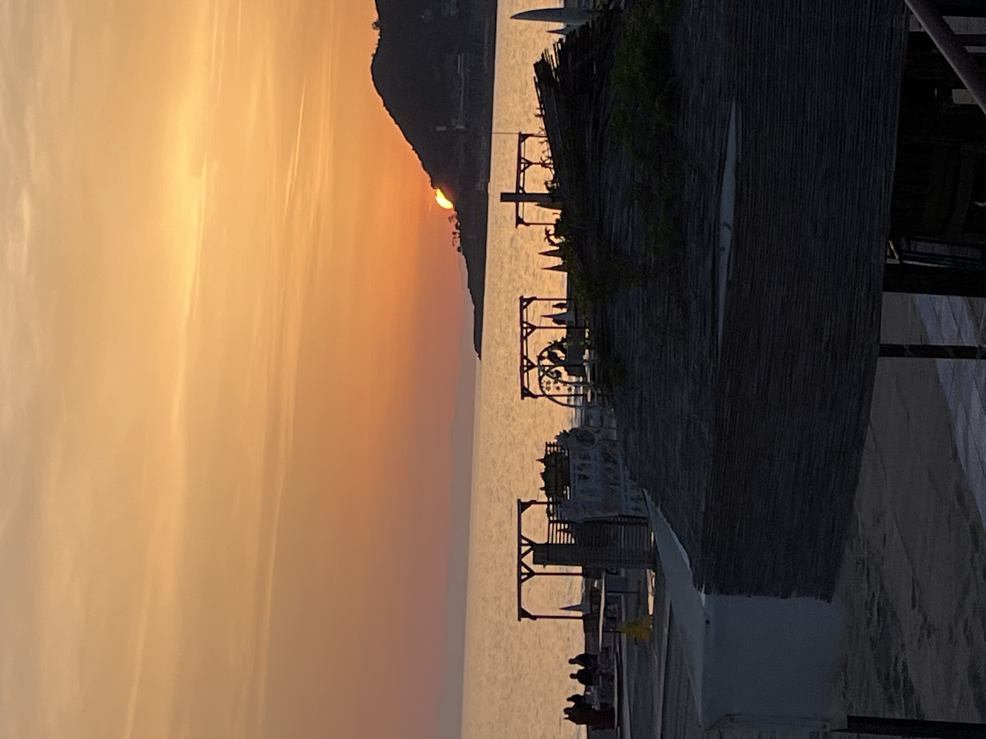 The image shows a coastal scene at sunset, likely in Alanya, Turkey, given the presence of a prominent headland. The sun is setting behind the headland, creating a warm, orange glow in the sky and reflecting off the water.
In the foreground, there's a structure, possibly part of a beach club or restaurant, with a textured wall and metal framework. People are gathered in the lower part of the image, suggesting they are at the beach or a seaside establishment. The lighting is dim due to the sunset.
