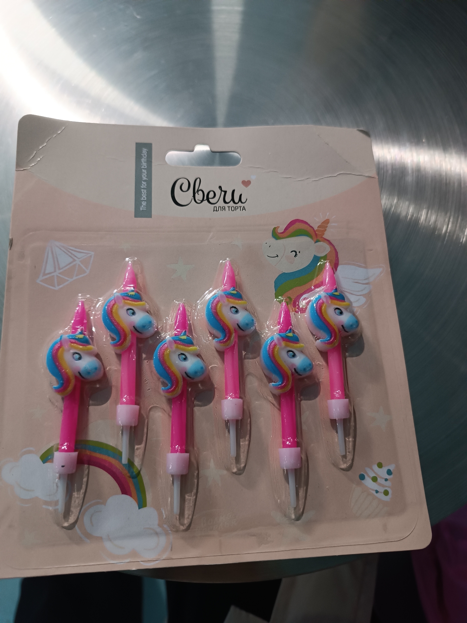 The image displays a retail package of six unicorn-themed birthday candles, positioned on a brushed metallic surface, likely a countertop, with ambient light reflections. The package consists of a light pink/beige cardboard backing with a clear plastic blister containing the candles. The cardboard features Cyrillic text 'Свечи' (Candles) and 'ДЛЯ ТОРТА' (FOR CAKE), and English text 'The best for your birthday'. Decorative elements include a cartoon unicorn head with a rainbow mane, a rainbow arch with clouds, a cupcake outline with sprinkles, and a faceted gem outline. The top left corner of the cardboard shows minor creasing. Each of the six candles is pink, approximately 7-8 cm long, and topped with a molded unicorn head figurine detailed with a light blue face, white muzzle, black eyes, pink horn, and a rainbow-colored mane. Each candle has a white plastic spike for insertion. Blurred portions of a teal object and dark purple fabric are visible in the lower corners of the frame. No individuals or specific actions are depicted. The location is Unknown, Unknown.