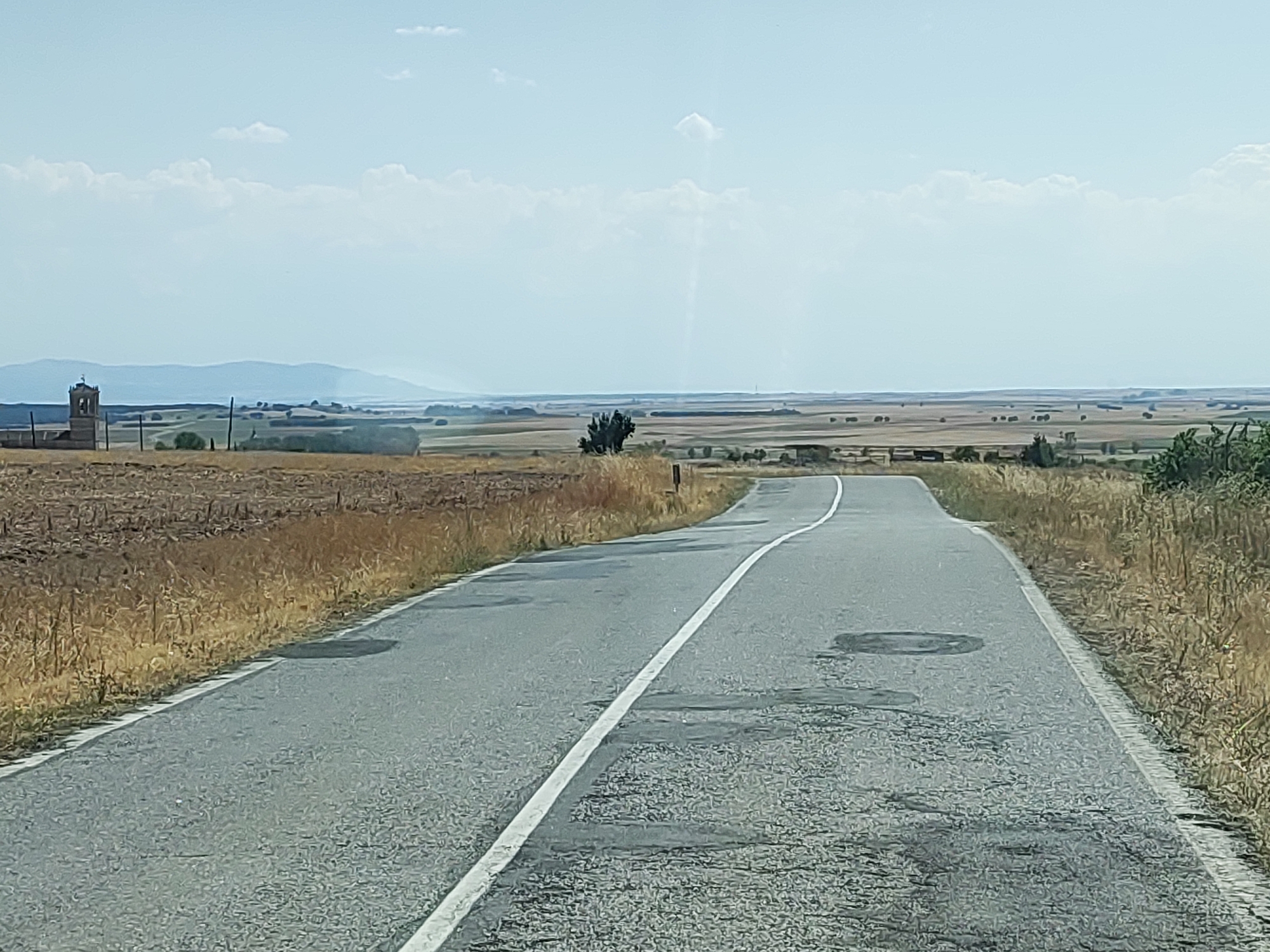 The image depicts a rural road curving through a dry, grassy landscape. On the left side of the road, a field of harvested crops or dry vegetation stretches towards a distant horizon. A weathered stone structure, possibly a bell tower or part of a church, stands in the far left of the frame, near the edge of the field. Scattered trees and some structures are visible in the mid-ground, appearing as small dots against the expansive, hazy landscape. The road itself is asphalt, marked with a white line down the center and showing signs of wear, with several patches and circular marks on its surface. The right side of the road is bordered by dry, yellowish grass. The sky is bright and clear, with a few scattered clouds and a faint vertical streak that might be a lens flare or atmospheric phenomenon. The overall impression is one of a hot, dry day in a sparsely populated rural area. The location is identified as being near Santa María la Real de Nieva, Spain, a municipality in the province of Segovia.