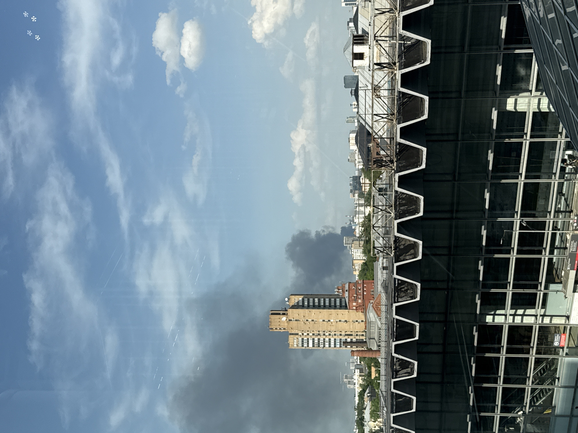 This image shows a section of Moscow, Russia, with a view of the sky, buildings, and reflective surfaces. The sky is blue with scattered clouds. On the right side of the frame, there is a dark, reflective facade of a building, its surface structured with a grid-like pattern. Adjacent to this, there's exposed metal scaffolding. Further into the distance, multiple buildings of varying heights and colors are visible. The weather seems to be changing, with a dark, cloud-filled area contrasted against the brighter blue sky. The overall orientation of the image is rotated 90 degrees.