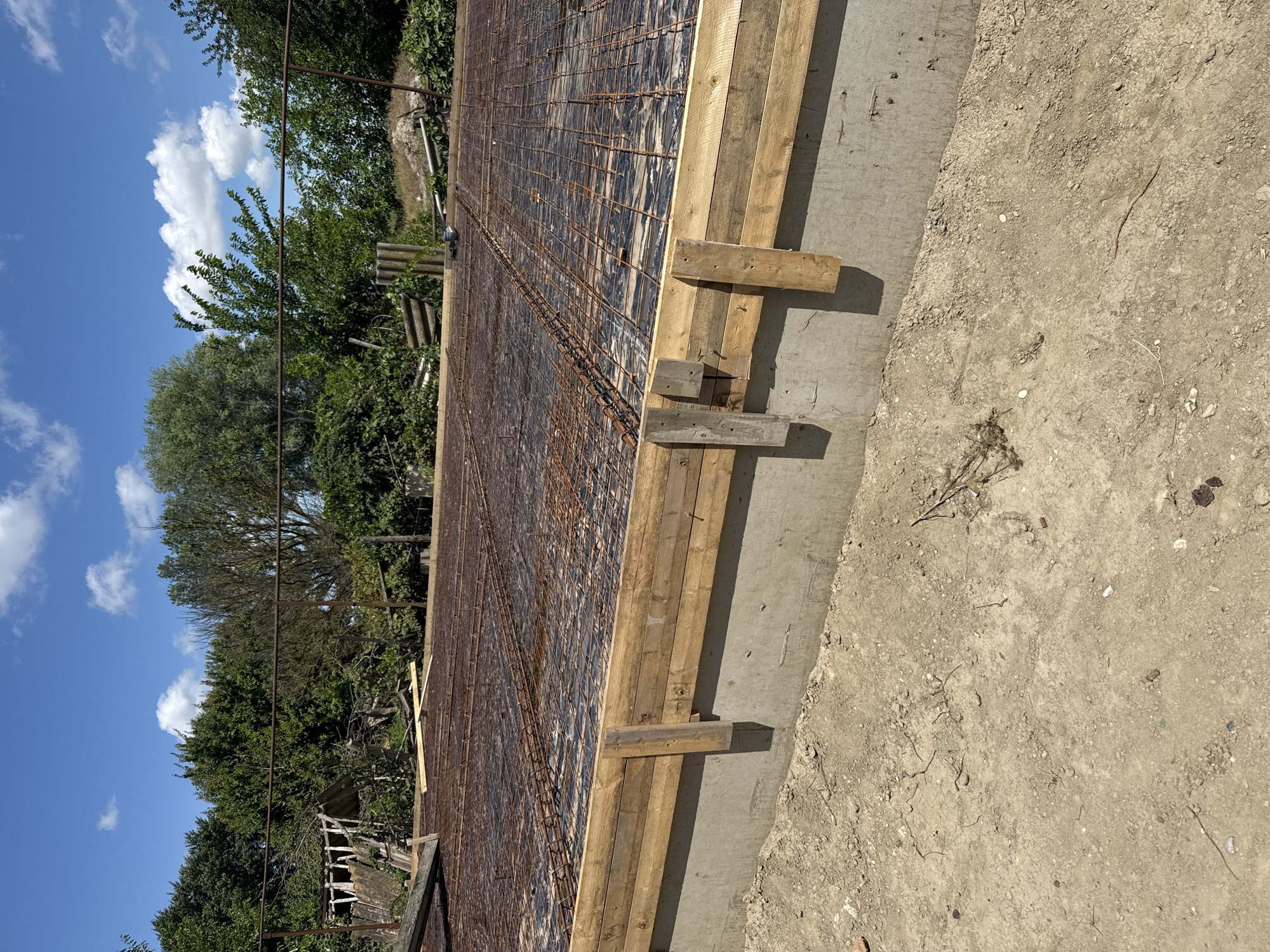 The image depicts a construction site, likely the foundation of a building, located outdoors in Yasnaya Polyana, Russia. In the foreground, dry, sandy soil with scattered small stones and debris covers the ground. A concrete foundation wall is visible, topped with wooden formwork. Inside the formwork, a layer of black plastic sheeting is laid down, on top of which a grid of rusty steel rebar is arranged, indicating preparations for a concrete pour. In the background, a dense line of various green trees and foliage forms the natural environment. A rustic metal fence runs horizontally across the scene, separating the construction area from the trees. The sky above is bright blue with scattered white cumulus clouds. No people are present in the image, and the scene is static, showing a stage of construction.