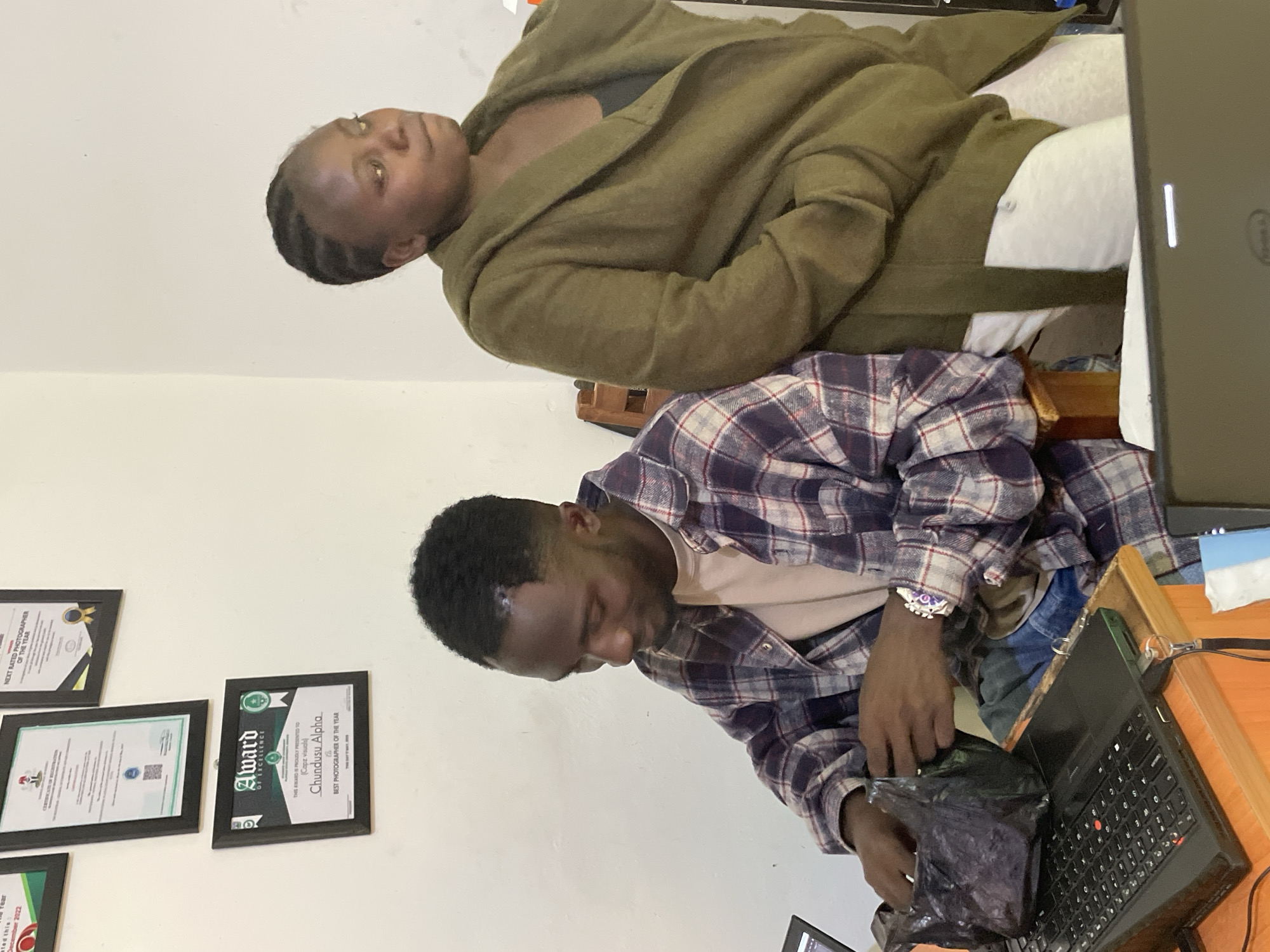 The image depicts an indoor scene, likely an office space in Durumi, Nigeria, as indicated by the presence of framed awards on the wall. In the foreground, a man wearing a plaid shirt is seated at a desk, focused on a laptop and handling a black plastic bag. A woman, dressed in a long olive-green coat, stands behind him, looking away from the camera. The environment suggests a working or administrative setting, with diplomas or certificates displayed on the wall.