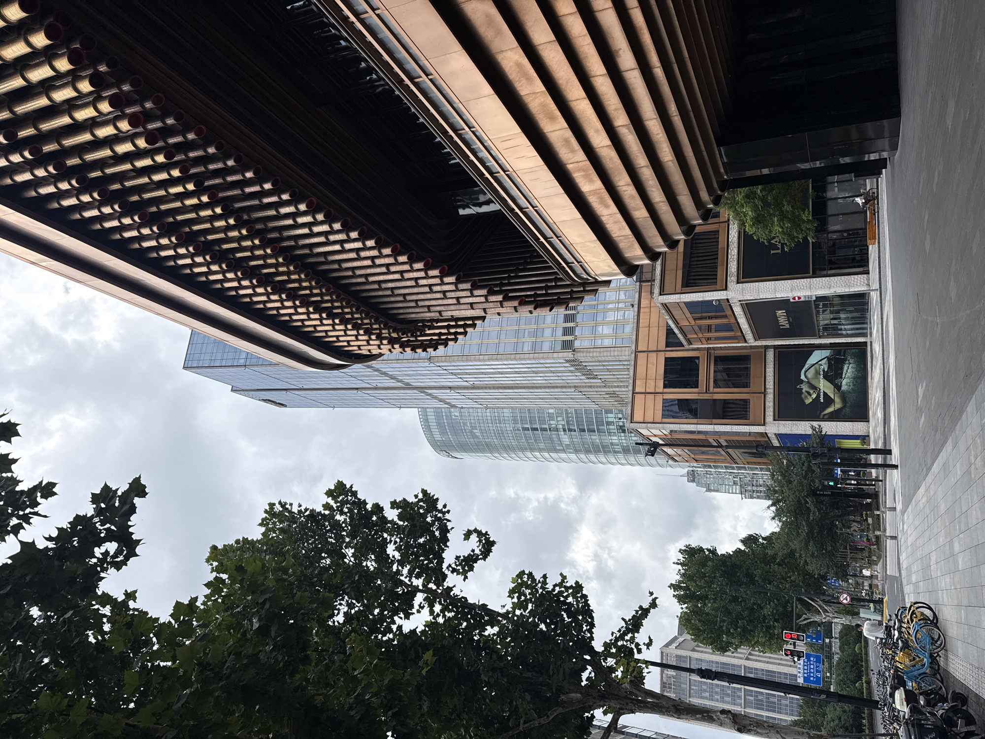 The image shows a street scene in Shanghai, China, on a cloudy day. In the foreground on the right is a modern building with a distinctive copper-colored facade featuring horizontal layers and vertical cylindrical elements. To the left, a tree with lush green foliage partially obscures the view of the sky. Beyond the copper building, a tall, sleek skyscraper with a glass facade rises into the sky. Further to the left, another modern building with a glass exterior is visible, and behind it, a rounded glass skyscraper stands out. In the middle ground, a street runs horizontally, lined with trees. On the left side of the street, several bicycles are parked. Traffic lights are visible above the street, with red lights illuminated. A pedestrian crossing is present on the sidewalk. A person on a bicycle is seen in the middle distance, riding away from the camera. Storefronts are visible on the ground floor of some buildings, with signage including "Alexander Wang" and "Lanvin." The overall impression is of a contemporary urban environment in Shanghai.