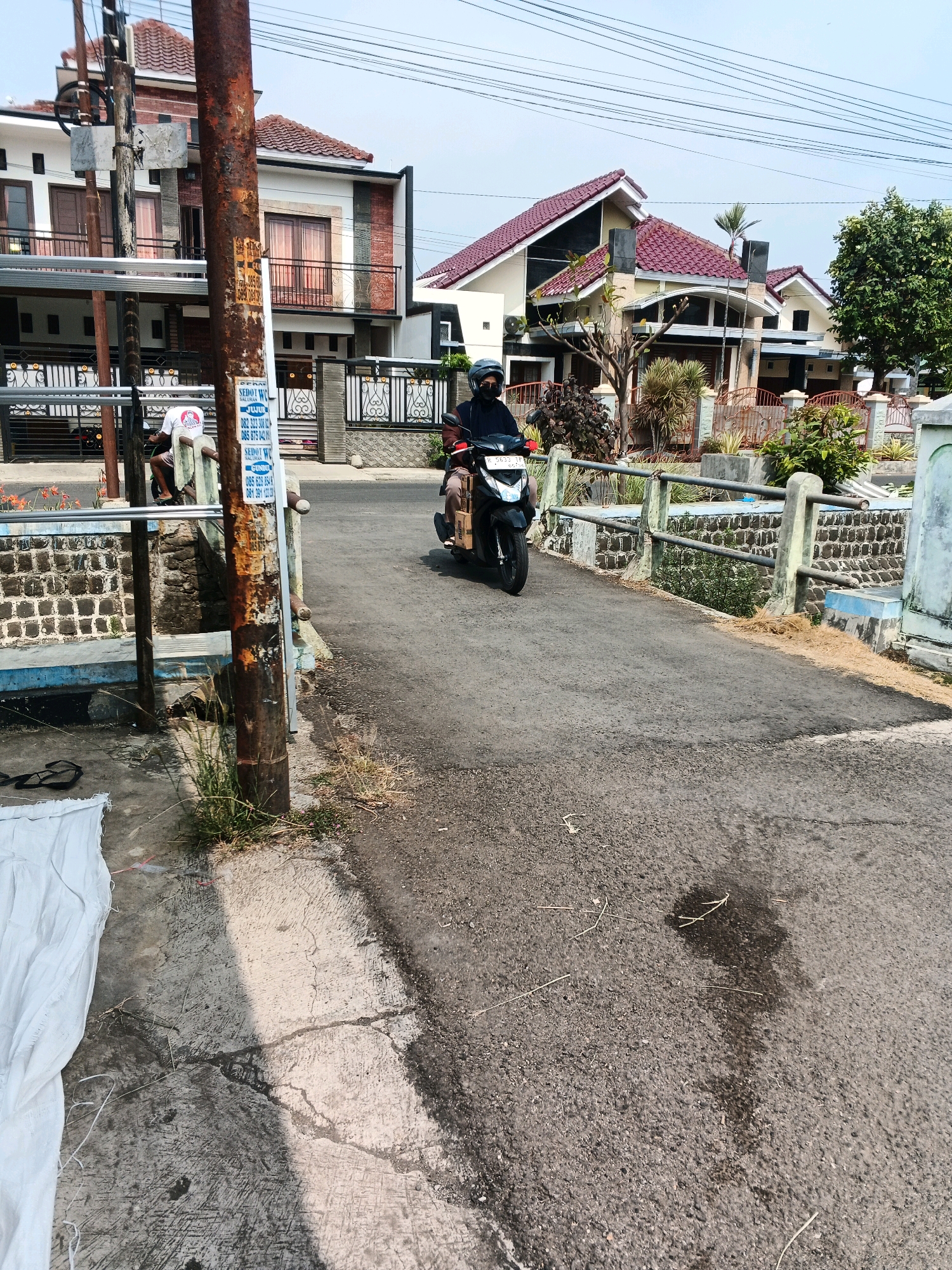 The image depicts a residential street in Kelurahan Tegalkamulyan, Cilacap, Indonesia. A person is riding a black motorcycle on an asphalt road. The rider is wearing a helmet and a dark jacket. On the left side of the road, there is a concrete sidewalk with some white fabric and grass. A rusty metal pole with a sign is also present on the left. In the background, there are several houses with red-tiled roofs and balconies. A stone wall with a metal railing runs along the right side of the road, separating it from a lower-lying area with vegetation. The sky is clear and bright.