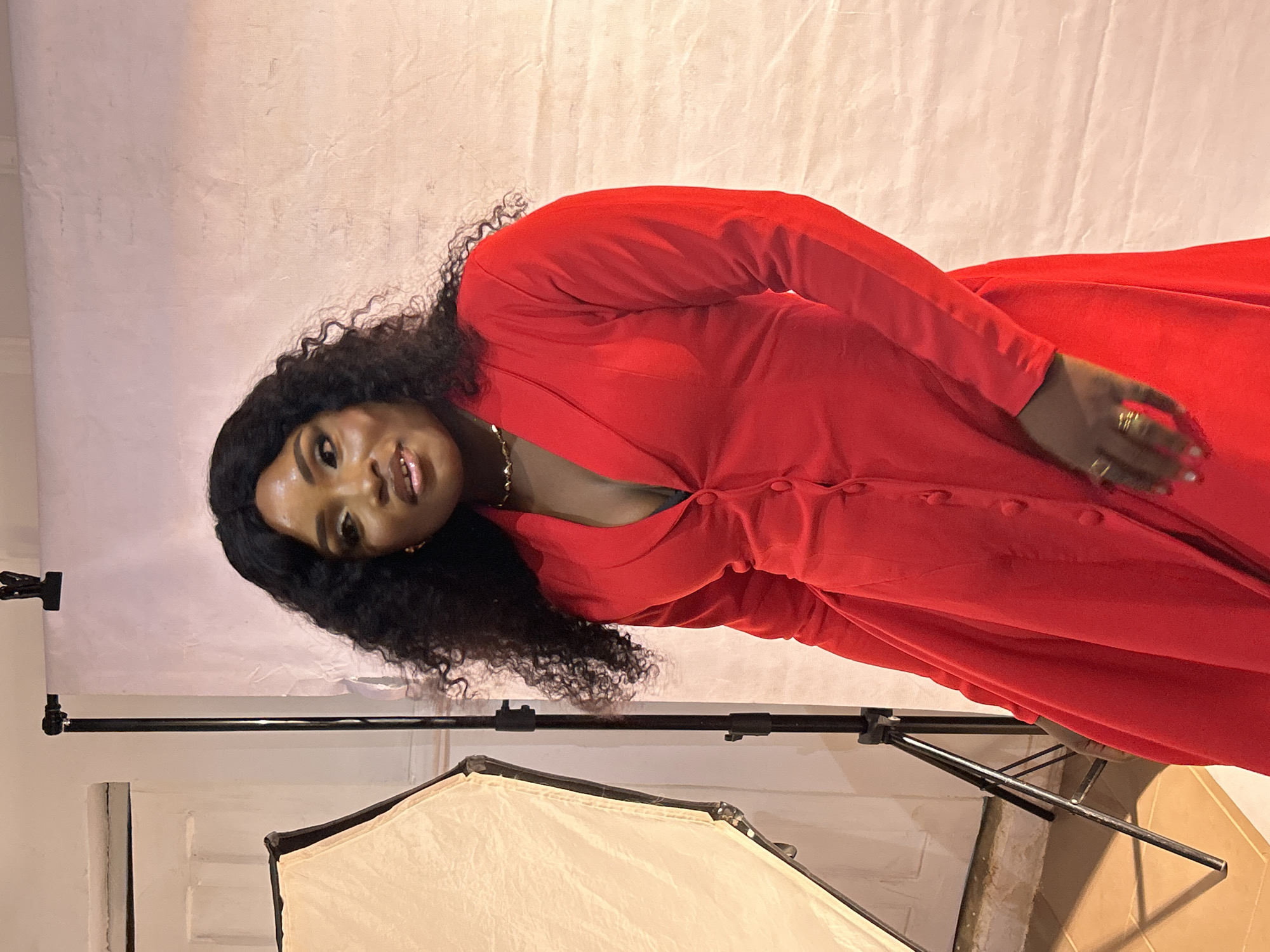 A medium shot depicts a woman posing in a red dress in what appears to be a studio setting, possibly in Jos, Nigeria. She has dark hair, and the dress has long sleeves and buttons down the front. A light source with a diffuser is visible in the foreground, and a backdrop is positioned behind her, held up by a stand. The scene suggests a professional photoshoot or a casual portrait setting.