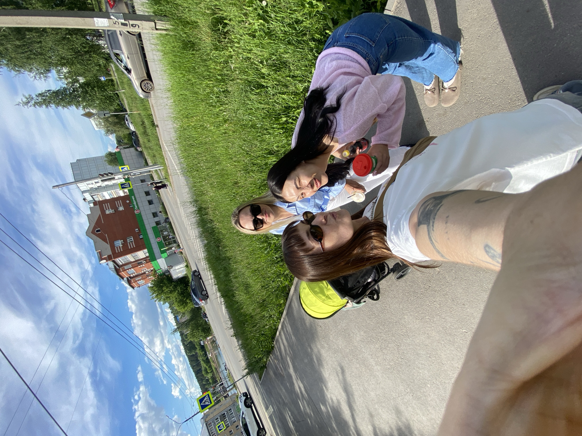 The image captures three women on a sunny day, positioned along a sidewalk in what appears to be an urban or suburban setting. The photograph is taken from a low, close-up perspective, possibly a selfie, looking upwards at the subjects and the surrounding environment.
In the foreground, a woman with brown hair, wearing sunglasses and a white top, is partially visible, facing away from the camera. Beside her, another woman with blonde hair and sunglasses is also seen from the side, wearing a light blue top. Further back, a third woman with long dark hair, dressed in a pink long-sleeved top and blue jeans, smiles broadly towards the camera. She holds a red and green cup along with a dark bottle, possibly a drink. A yellow and black handle, likely from a stroller or pram, is visible near the foreground woman.
The setting includes a paved sidewalk alongside a strip of vibrant green grass, which borders a road. Several cars are parked or driving on the road. In the background, there are multi-story buildings, traffic lights, and various street signs, including a pedestrian crossing sign. One building is a reddish-brown brick structure, while another features prominent green signage. Visible text on these signs includes "Fix Price" and a partially visible "Улыбка радуги" (Ulybka Radugi), both indicating retail establishments. Overhead, power lines crisscross a bright blue sky dotted with scattered white clouds. The time of day is clearly daytime, evidenced by the bright sunlight and visible shadows. The overall mood conveyed is casual and cheerful, suggesting friends enjoying an outdoor outing in Votkinsk, Russia.