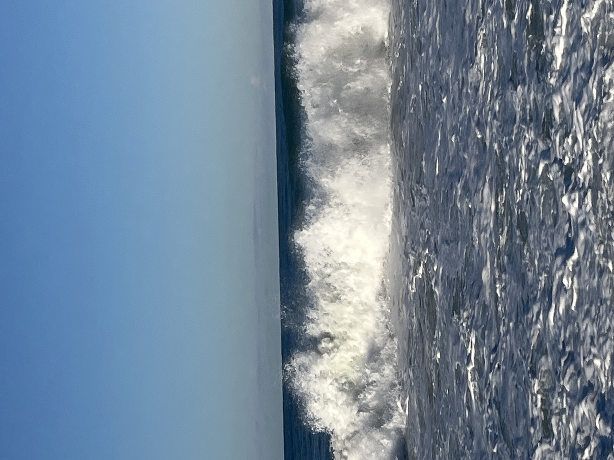 The image shows a wave crashing on the shore near Alanya, Turkey. The sky is a clear, light blue. The wave is large and white, with foamy crests. The water is a dark blue with white caps. The image appears to be taken at a low angle.