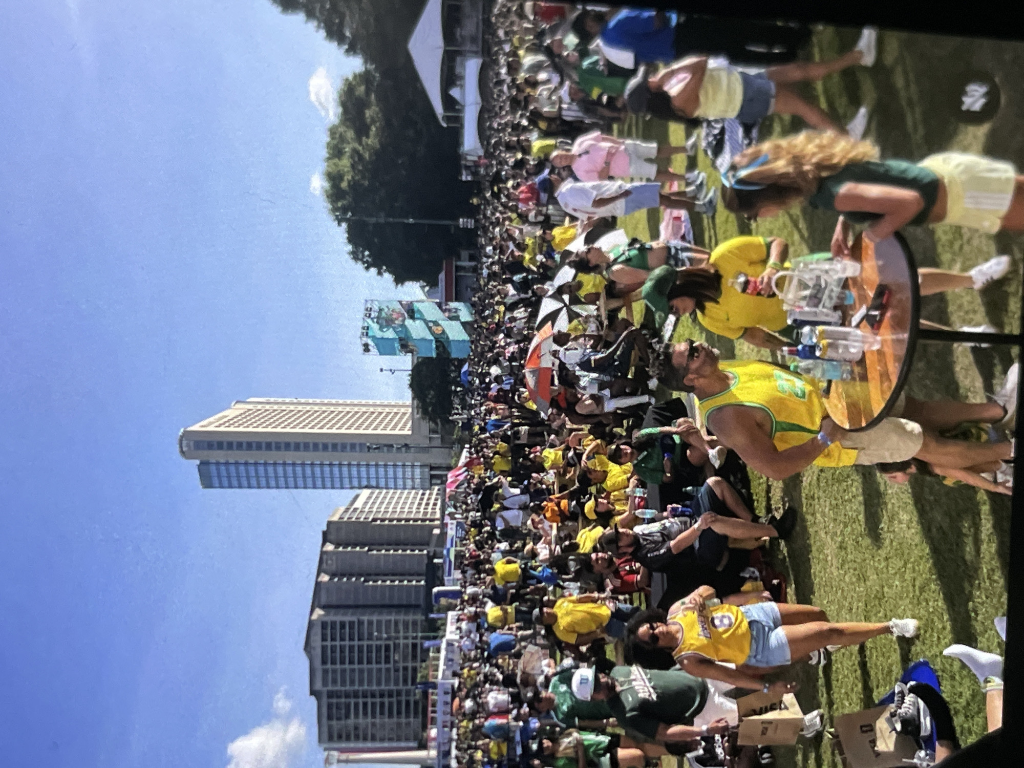 In a sunny, outdoor urban environment, a large crowd gathers on a grassy field in front of several tall buildings. Many people are dressed in yellow and green, suggesting a possible sporting event or celebration. Some are seated, while others stand and interact. In the foreground, a round table is set up with bottles and a few people are gathered around it. The scene appears lively and festive, with a mix of genders and ages. The backdrop includes a clear blue sky with scattered clouds and a line of trees. A large, colorful structure with screens is visible in the distance, adding to the event atmosphere.