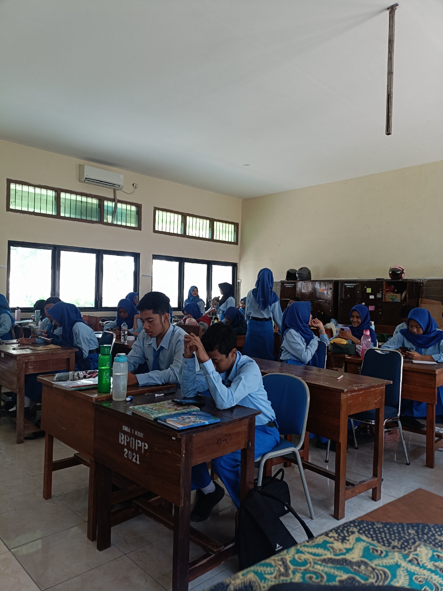 This is an indoor photograph of a classroom scene in Kediri City, Indonesia. The room appears to be a standard educational facility with rows of wooden desks and chairs. Several students, predominantly wearing blue uniforms and headscarves, are seated at their desks, engaged in various activities. Some are looking at books, others are using mobile phones, and a few appear to be resting their heads on their arms. Natural light streams in from large windows with metal gratings, and an air conditioning unit is mounted on the wall. The overall impression is that of a typical school day in session.