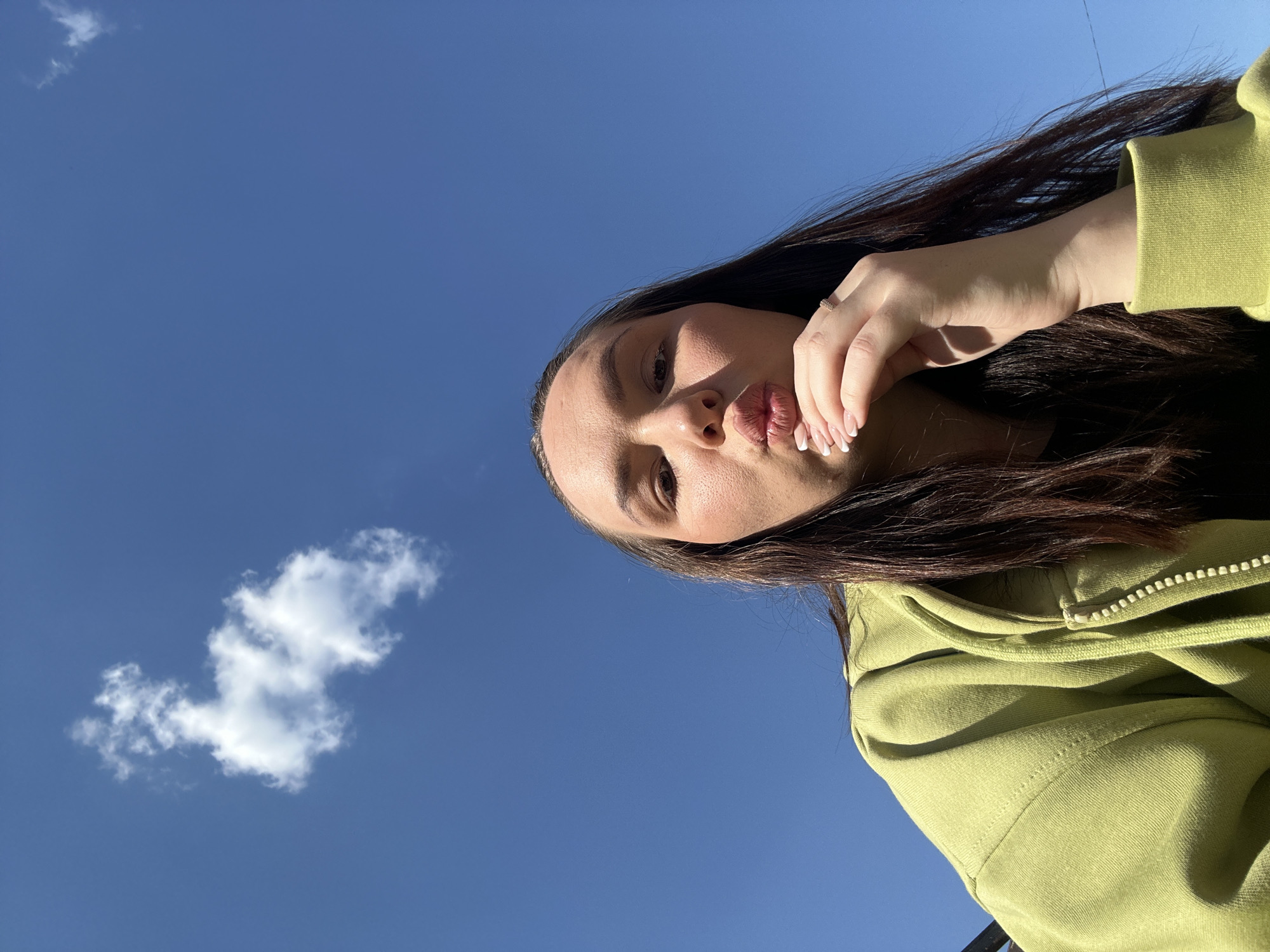 A low-angle, eye-level shot captures a woman against a clear, bright blue sky with a single white cloud in the lower left corner. The woman, positioned in the upper right portion of the frame, has long, dark brown hair. She is wearing a light green zippered hoodie. Her right hand is raised to her face, with her fingertips touching her cheek. She has manicured nails with white tips. She is pursing her lips into a kiss. The location is Chaikovsky, Russia.