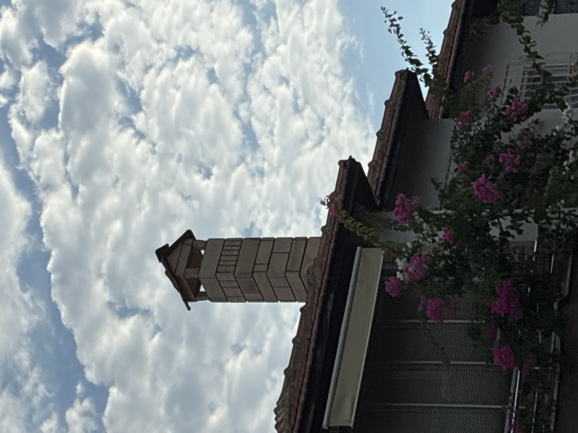 A chimney made of tan bricks stands tall against a sky filled with white, fluffy clouds. The chimney is topped with a dark brown, angular roof. Below the chimney, a brown tiled roof covers a building with a white facade. A section of the building has a retractable awning. Pink flowers bloom on vines growing up the side of the building, some reaching towards the chimney. The scene is likely in Alanya, Turkey, given the architectural style and vegetation.