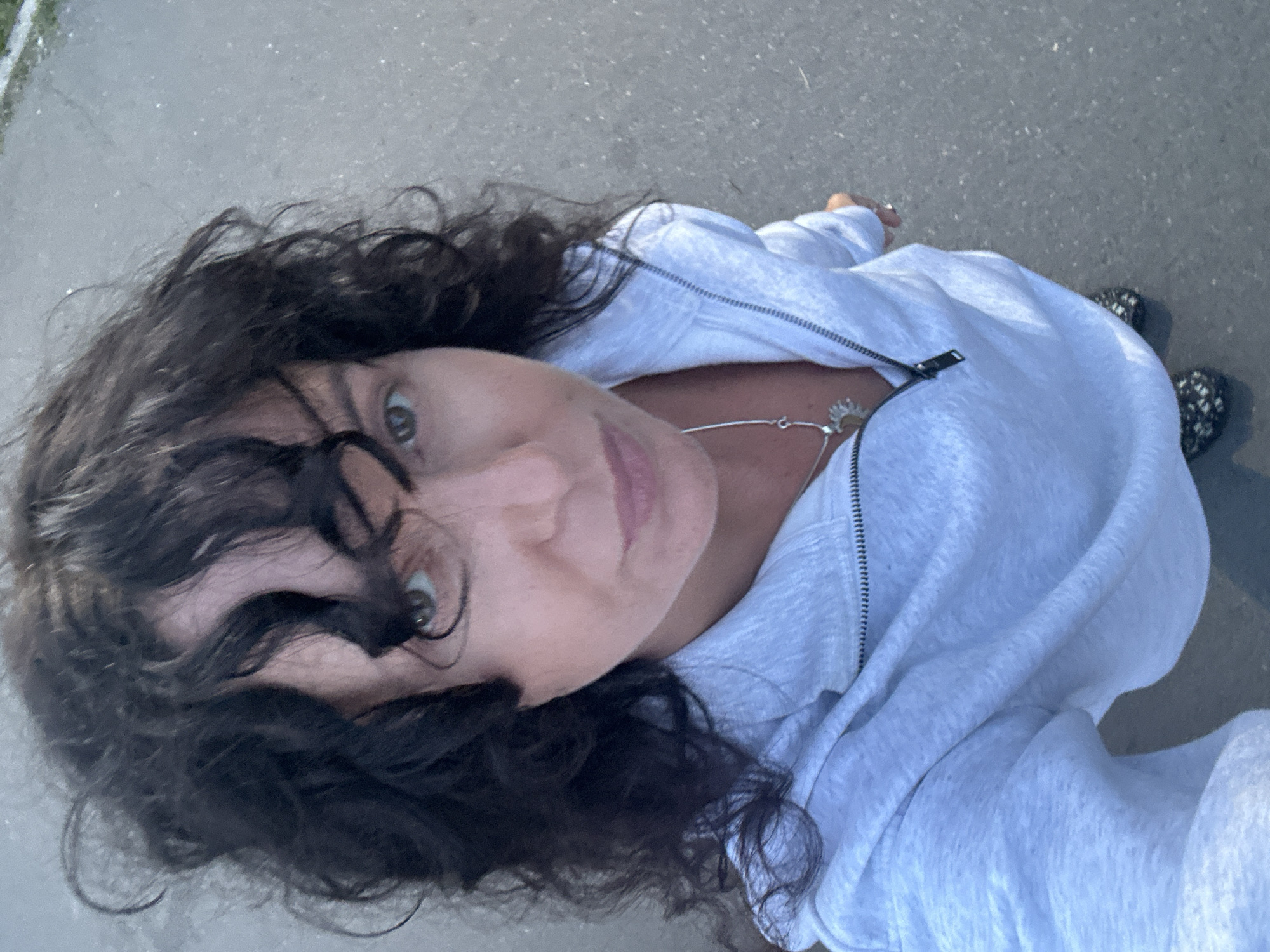 The image shows a person with dark, curly hair and green eyes, looking directly at the camera. They are wearing a light gray hooded sweatshirt and a silver necklace with a sun pendant. The sweatshirt is partially unzipped, revealing the necklace. The person is standing outdoors on an asphalt surface. The lighting suggests it is daytime, possibly late afternoon or early morning, with soft sunlight. The scene appears to be a casual, everyday setting, possibly a street or park. There are no visible signs of text in the image. The overall mood is relaxed and informal.