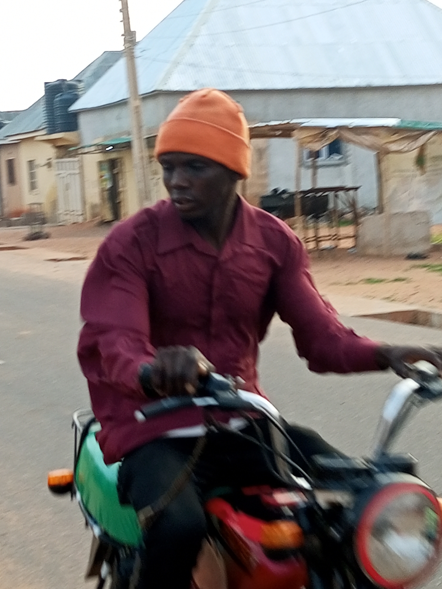 A man wearing a maroon long-sleeved shirt and an orange beanie is riding a motorcycle down a paved street in Tempure, Nigeria. The man is looking to his right, with his hands on the handlebars. The motorcycle has a green seat and a red headlight. In the background, there are buildings with metal roofs and some trees. The sky is overcast.