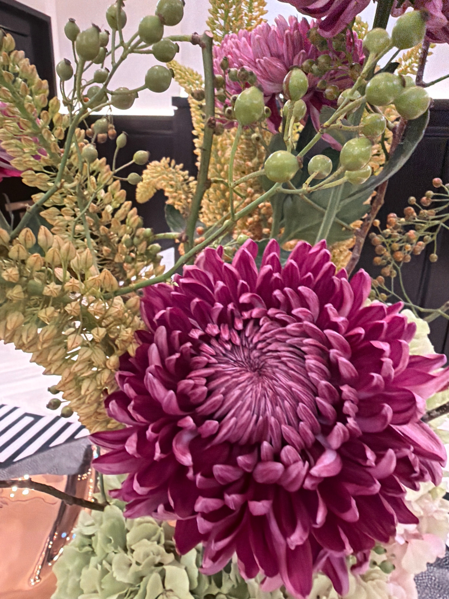 A close-up, eye-level, medium shot captures a vibrant floral arrangement. The primary subject is a large, deep pink mum with numerous delicate petals, its center tightly coiled. Behind and to the left of the mum, a spray of small, light green berries on thin stems adds a contrasting texture. Further left and slightly behind, a cluster of pale yellow, bud-like flowers on thin stems provides another layer of visual interest. A few small, dried brown seed heads are visible in the upper right background. The arrangement is set against a dark, out-of-focus background, with a hint of a striped pattern and what appears to be decorative lighting in the lower left corner. There are no people present, and no specific geographical landmarks are identifiable that would suggest a location in Moscow, Russia.