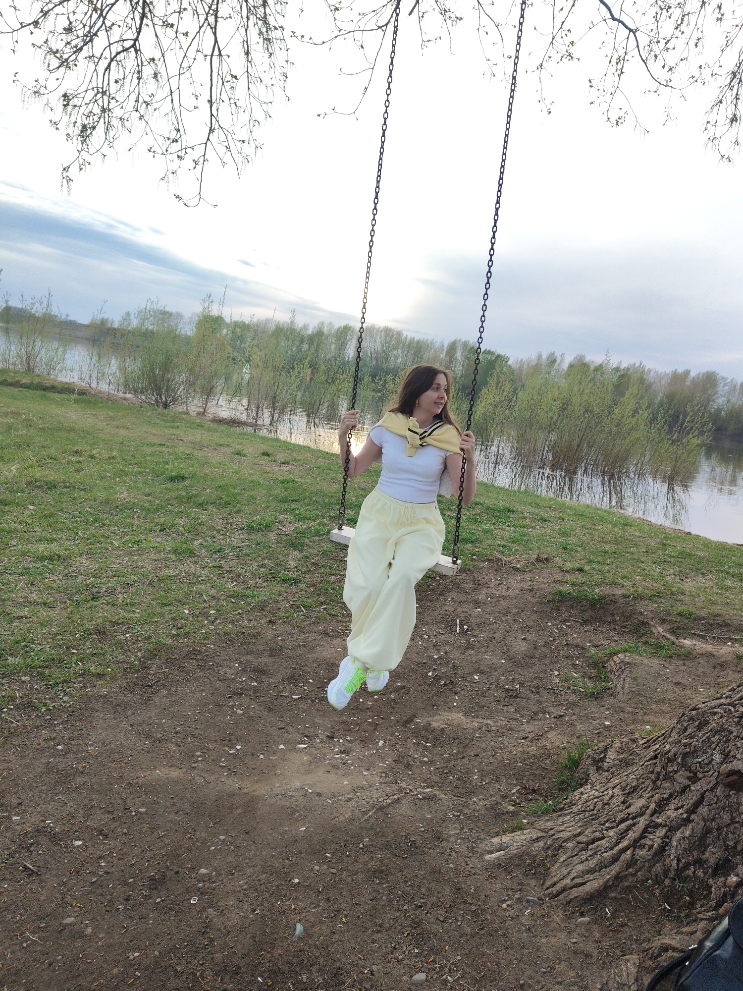 A young woman is sitting on a swing suspended from a tree. The swing is located in a grassy park with trees and a body of water in the background. The woman is wearing a white shirt, light yellow pants, and sneakers. She has a yellow sweater draped over her shoulders. The location is Unknown, Unknown.