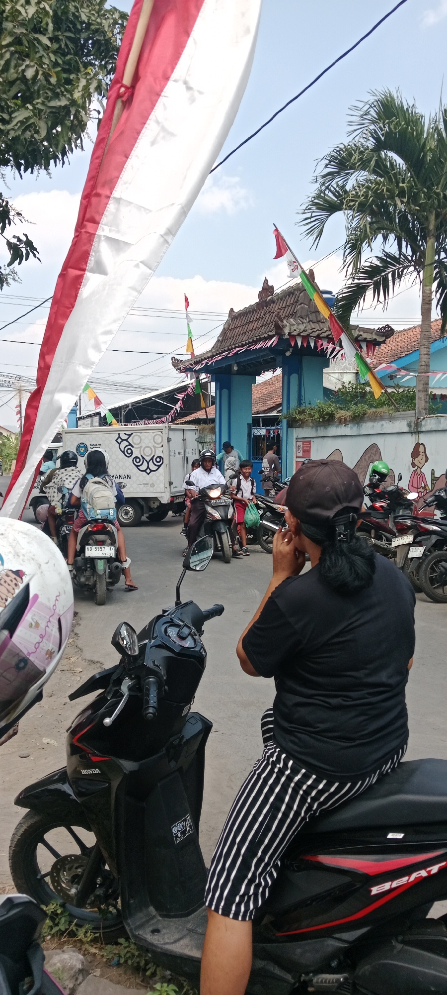 This is a street scene in Purwomartani, Indonesia, likely a residential area given the presence of a school or community building. In the foreground, a person wearing a black t-shirt and black and white striped pants is sitting on a black scooter, with their back to the camera, and appears to be touching their ear. Another scooter is partially visible in the lower left, with a helmet on it.
In the middle ground, traffic is present. Several people are on motorcycles. One motorcycle carries a driver and a passenger wearing a backpack. A white truck with graphics on its side is also visible, and a man in a white shirt is riding a motorcycle. Two young boys in school uniforms are also on a motorcycle. The entrance to a blue building with a traditional tiled roof, decorated with red and white flags and colorful pennants, is prominent in the background. Palm trees are visible behind the building. The sky is blue with some clouds. The overall atmosphere suggests a bustling street, possibly during a local celebration or school day.