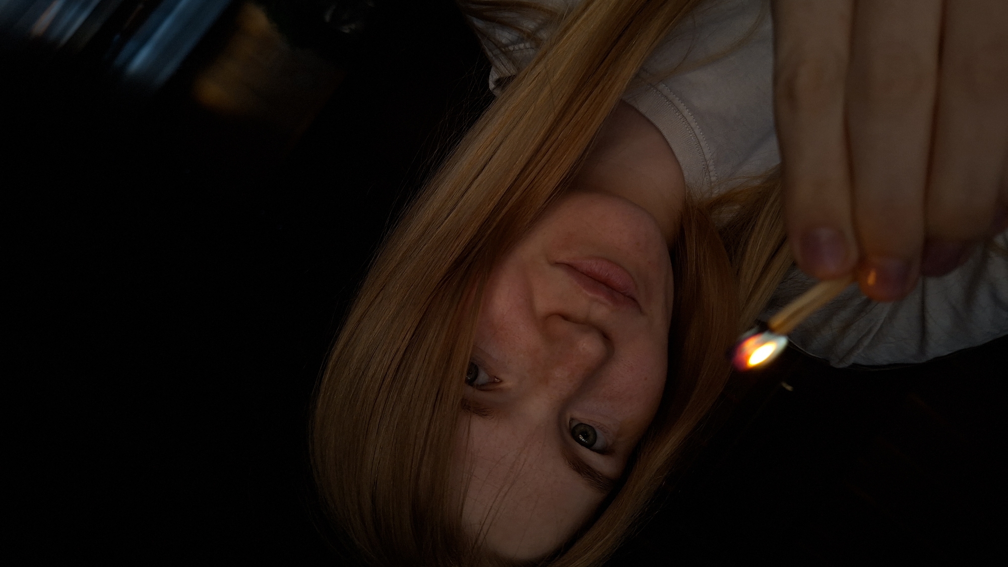 A young woman with long, light brown hair and freckles is holding a lit match in her right hand. She is looking towards the camera with a slight smile. The background is dark.