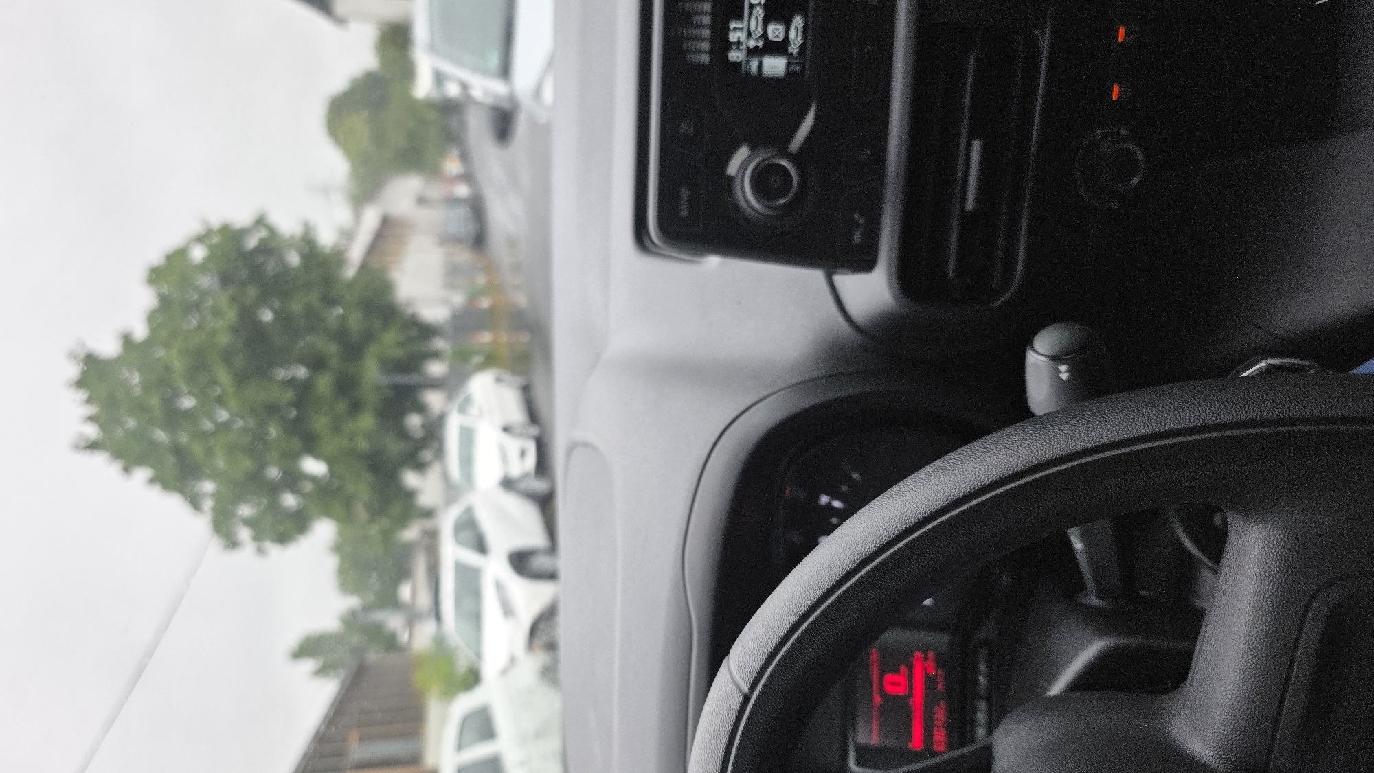 The image is taken from inside a car, looking forward through the windshield. The dashboard is visible in the foreground, with a steering wheel on the left and a car radio on the right. The radio display shows the time as 8:51. Outside the car, it appears to be a cloudy day. There are several white cars parked along a street, and a large green tree is visible in the background. Buildings line the street on either side, and the ground appears wet, suggesting it may have recently rained. The scene is an urban environment, possibly a residential street or a commercial area. There are no people visible in the image.