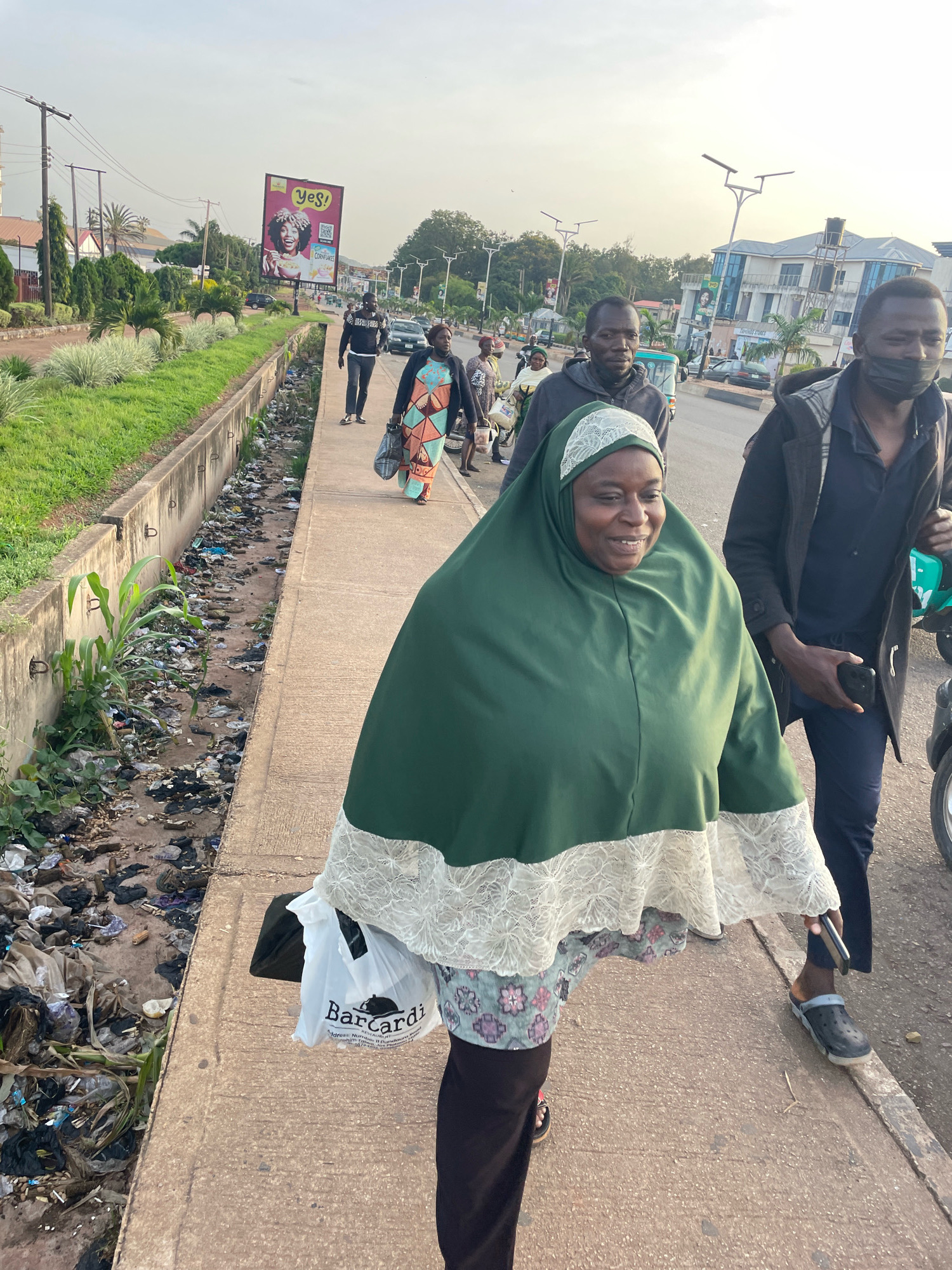 In Barakin Lerawi, Nigeria, a group of people walk along a sidewalk next to a street. The sidewalk is concrete and runs alongside a drainage ditch filled with refuse. On the street, a man in a black jacket and dark pants walks with a black bag in his hand. Next to him, a woman wearing a green headscarf and a patterned dress is walking, holding a white plastic bag with "Barcardi" printed on it. Behind them, another man in a black hoodie walks, and further back, a man in a black jacket and dark pants with a black face mask is seen. In the background, trees and buildings line the street, and a billboard with an advertisement is visible. The sky is overcast.