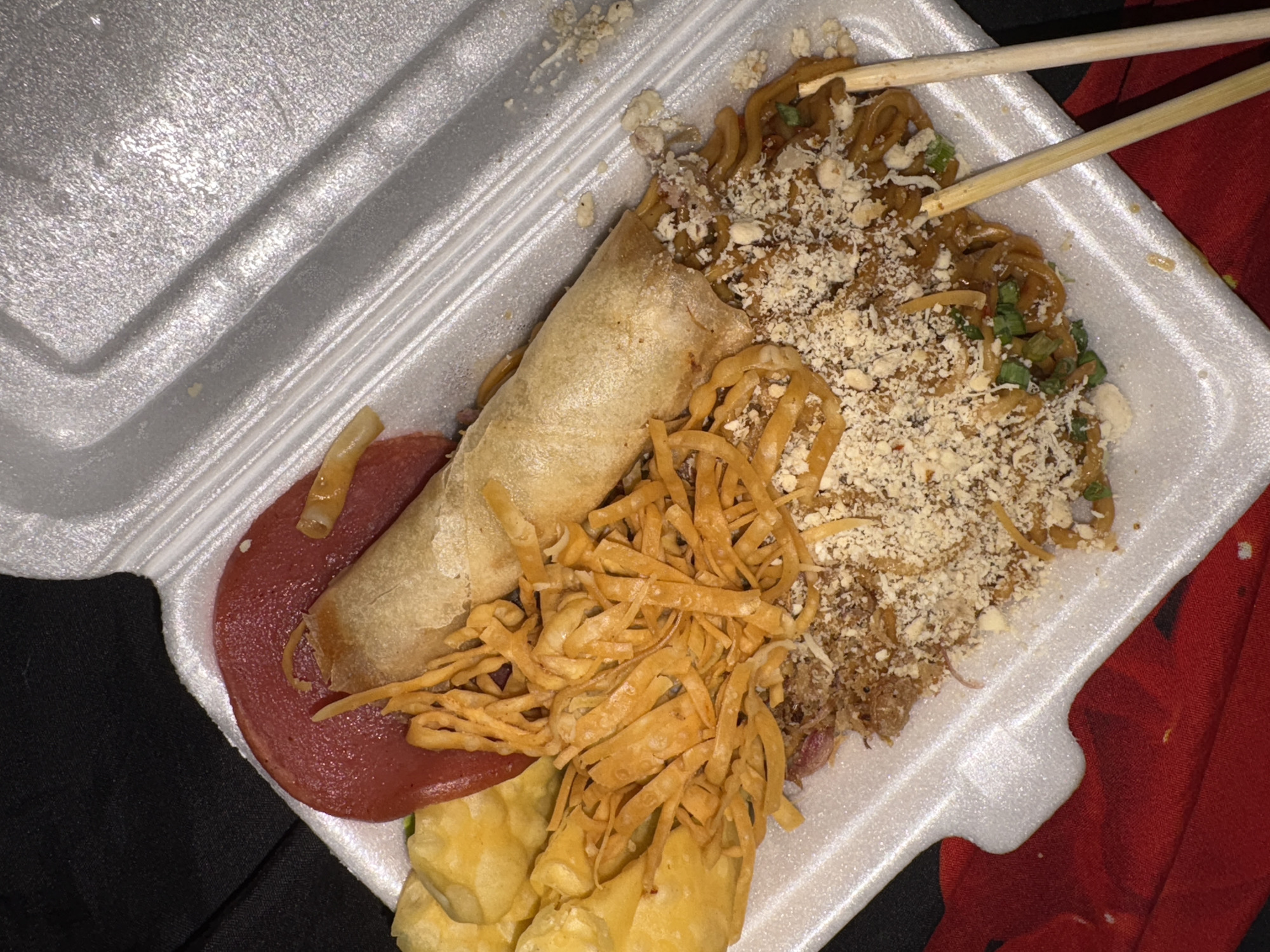 A close-up, overhead shot shows a to-go container filled with Asian-inspired food. The container is white styrofoam with raised ridges. The food is densely packed, obscuring the bottom of the container.
Dominating the center of the image is a golden-brown spring roll. Partially resting on the spring roll is a thin, circular slice of red meat, possibly ham or bologna. To the left of the spring roll, a pile of crispy, orange-colored fried noodles is visible. Beneath the spring roll and extending to the right are dark brown noodles mixed with green scallions and a topping of crushed nuts or breadcrumbs. Two wooden chopsticks lie horizontally across the noodles on the right side of the frame.
In the upper left corner, several golden-yellow fried wontons or dumplings are partially visible. The background is dark and out of focus, with hints of black fabric on the left and a red textured fabric on the lower right.
The image quality is moderate, with some graininess and slightly oversaturated colors. The lighting appears to be artificial, casting subtle shadows within the container. The scene suggests a casual meal, likely purchased for takeout or delivery.
The requested location of Gadingrejo, Indonesia, is not visually discernible from the image.