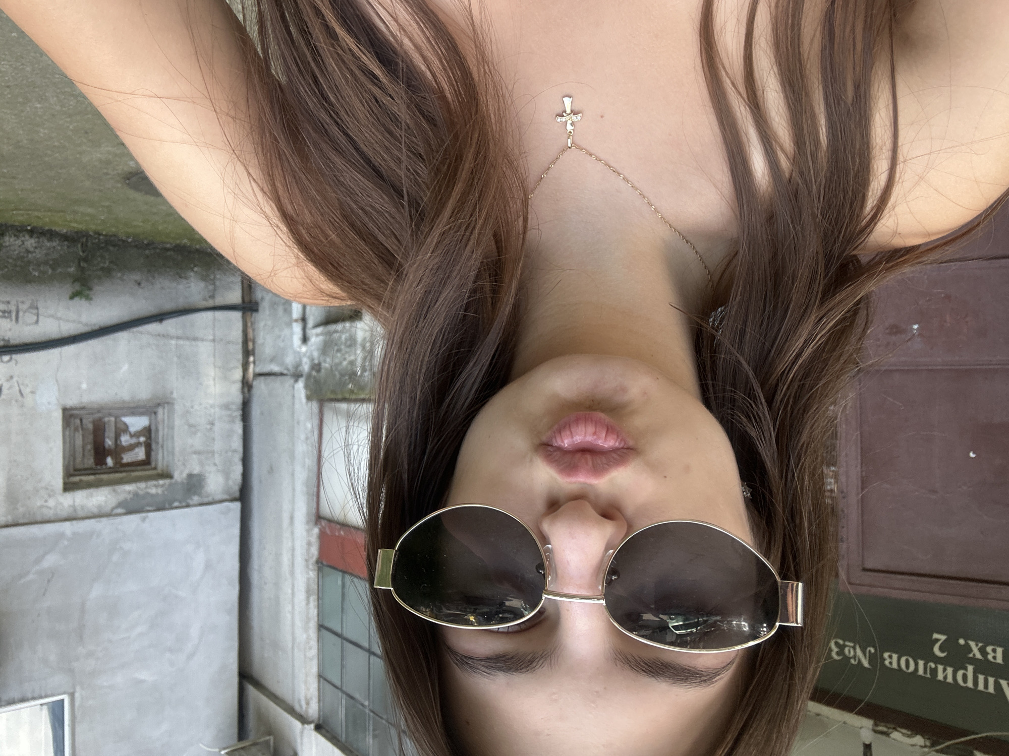 In Shumen, Bulgaria, a young woman with long brown hair, wearing sunglasses and a gold cross necklace, poses for a selfie. Her lips are pursed. She is standing in front of a building with dark brown doors and green signage that reads "Априлов №3 BH. 2". The background consists of a grey concrete building with windows. The overall setting appears to be an urban environment.