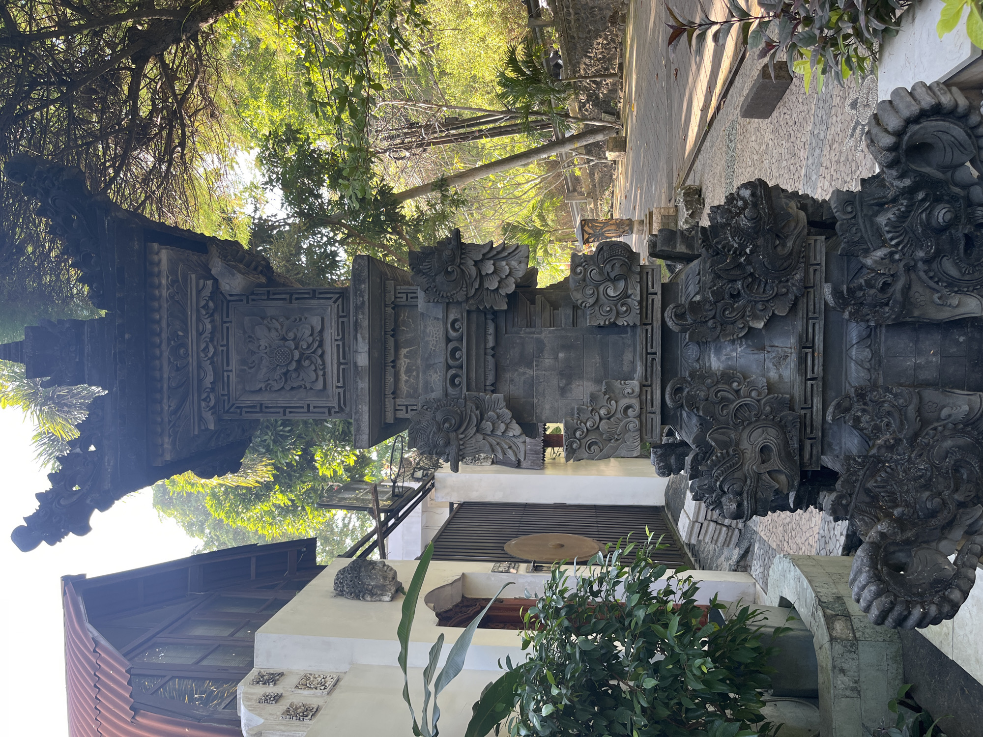 In Kuta Selatan, Indonesia, a stone shrine with intricate carvings stands in a paved outdoor area. The shrine features multiple levels, each adorned with decorative elements and stylized dragon-like figures. To the left of the shrine, a white wall is visible with decorative embossed patterns and a stone head sculpture mounted on it. A large green plant with broad leaves grows in a planter to the left of the wall. Beyond the wall, a wooden door and a metal gate are present. A building with a red corrugated metal roof and wooden-framed windows is situated to the left of the shrine. The background shows a pathway or road surrounded by trees and lush greenery, indicating a tropical environment. The lighting suggests it is daytime, with sunlight filtering through the trees.