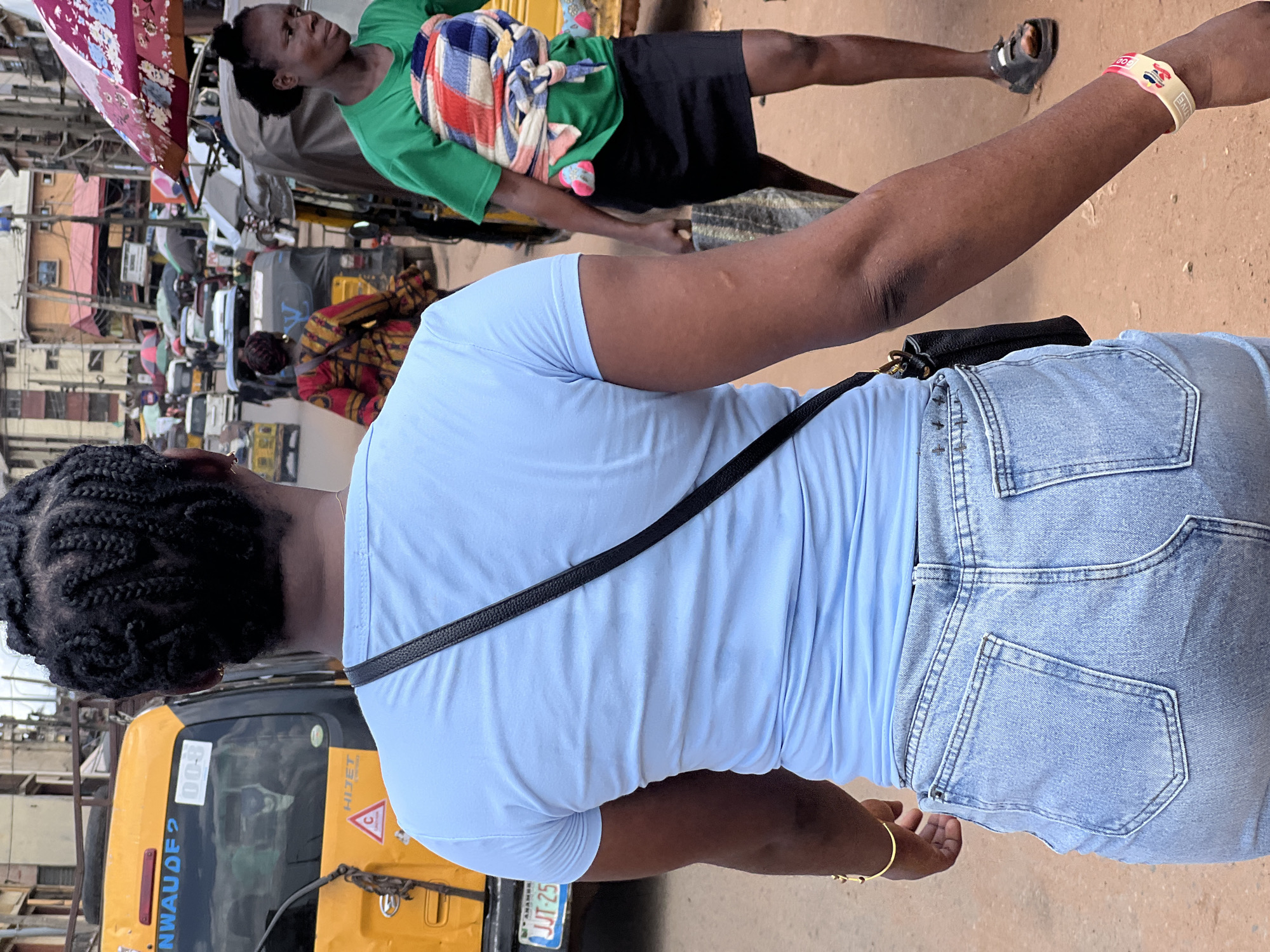 The image captures a street scene in an unidentified city. The foreground shows a person with braided hair, wearing a light blue shirt and light blue denim shorts with a black shoulder bag. In the background, a yellow auto-rickshaw or "Keke Napep" vehicle is partially visible. There are other individuals and vehicles suggesting a bustling street environment. One person is seen wearing a green shirt, black shorts and a colorful garment, while holding their arm with a wristband. The background shows various buildings and overhead electrical lines typical of an urban setting.