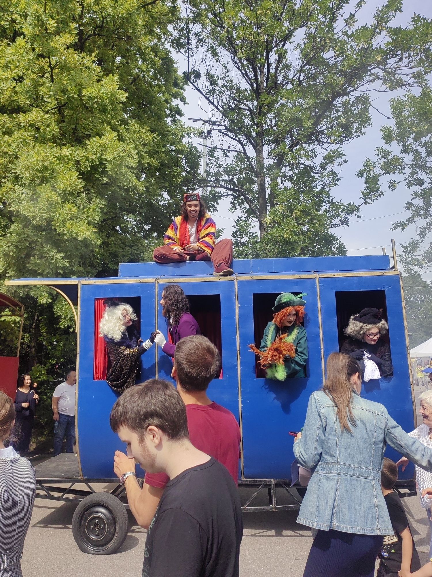 The image depicts an outdoor event with a blue stage structure, resembling a train car, as the central focus. Five individuals are visible on or around the stage. At the top of the structure, a person wearing a colorful, striped jacket and dark pants sits cross-legged, looking towards the camera with a smile. Below, in four windows of the blue structure, are four performers. The leftmost window features a person with blonde, curly hair, a dark, sparkly outfit, and a white glove. Next to them, a person with long, dark curly hair and a purple jacket is visible. The third window shows a person with bright red hair, a green hat, and a green outfit, holding a bundle of orange material. The rightmost window contains a person with gray curly hair, glasses, a dark outfit, and a black hat, holding a white cloth.
In front of the stage, several spectators are present. A young person with short brown hair is in the foreground, facing away from the camera and looking down. To their left, another person in a red shirt is partially visible. Further to the left, a woman in a dark dress and a man in a white t-shirt and jeans are standing. On the right side of the stage, a woman in a denim jacket and a dark skirt is holding a red object, and a child in a black shirt is standing nearby. An elderly woman in a striped shirt is also visible on the far right.
The environment appears to be a park or an open area with trees in the background and a paved surface in the foreground. The sky is visible and appears partly cloudy. The overall scene suggests a performance or a festival.
