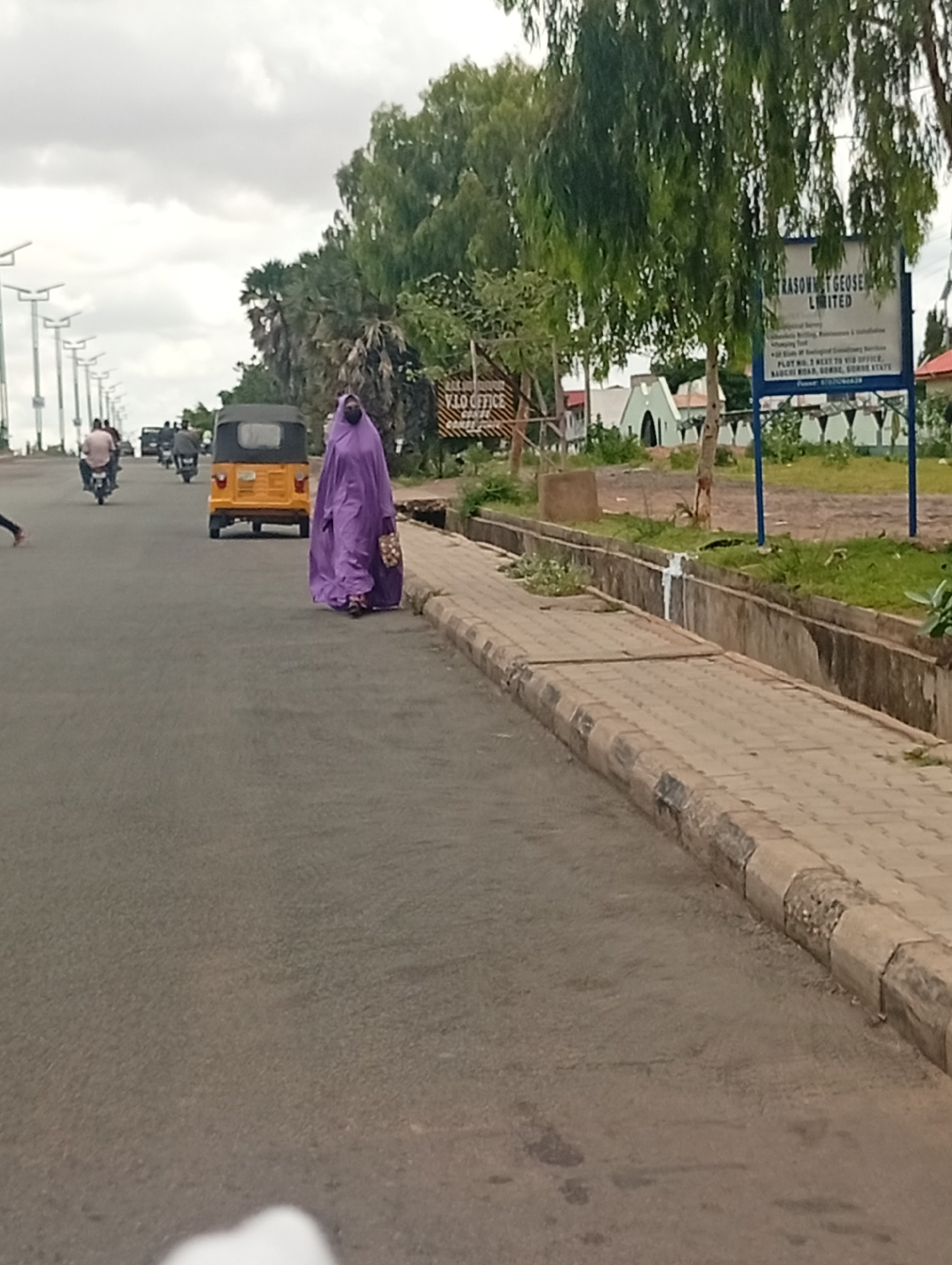 The image captures a street scene in Gombe, Nigeria, likely near a business district given the signage. A woman dressed in a vibrant purple hijab and abaya is walking on the right side of the asphalt road, carrying a patterned bag. She is wearing a face mask. To her left, a yellow auto-rickshaw is parked. In the background, several motorcycles are visible on the road, with riders, and a person is walking on the far left of the image. Tall trees with green foliage line the side of the road, and a concrete sidewalk with a drainage channel is present on the right. A blue and white sign for "ULTRAVISION GEOSERVE LIMITED" is prominently displayed, indicating services like "Geological Survey, Reservoir & Installation," "Well Logging & Testing," and "Oil & Gas Consulting Services." Further down the road, another sign reads "YAO OFFICE." The sky is overcast. The overall impression is that of a typical urban environment in Gombe, Nigeria, with mixed vehicular and pedestrian traffic.