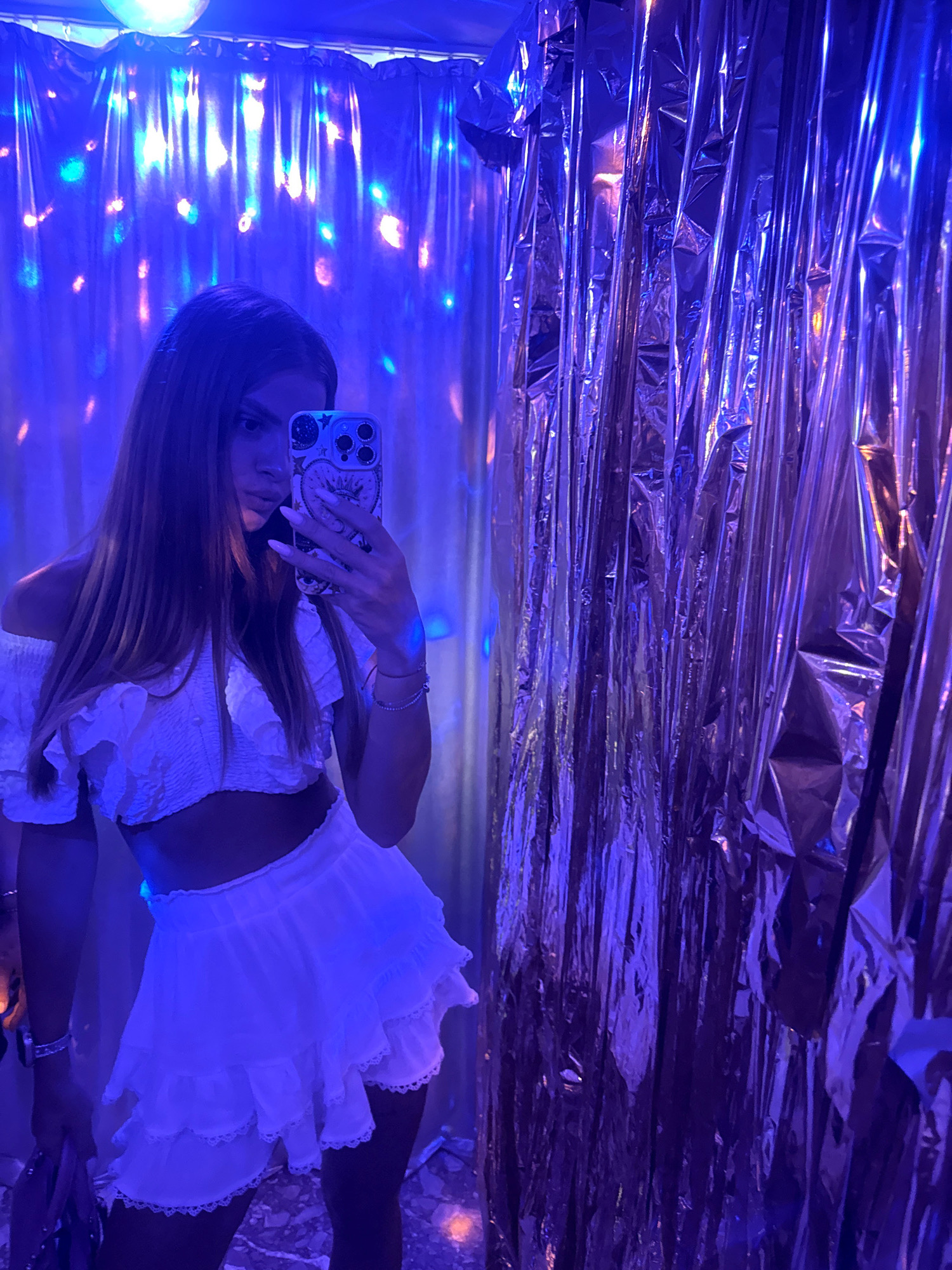 A young woman with long, dark hair is taking a selfie in a room decorated with blue and purple lights. She is wearing a white off-the-shoulder crop top with ruffles and a matching white ruffled skirt. She is holding a smartphone with a patterned case in her right hand. The background consists of shiny, crinkled metallic curtains and a ceiling adorned with lights. The overall atmosphere is festive and dimly lit. The specific location in Gabrovo, Bulgaria is not discernible from the image.