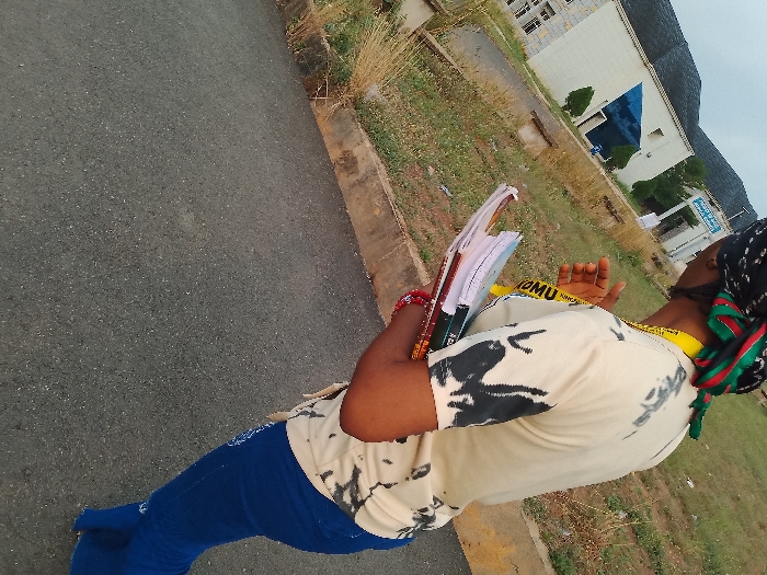 The image depicts an outdoor scene on the campus of the Federal Polytechnic, Ogboko, Nigeria. In the foreground, a dark-skinned female is observed from the mid-torso down, positioned at an angle as if walking. She wears a cream-colored short-sleeved t-shirt with black abstract patterns, blue pants, and a black-and-white patterned headscarf secured by a green and red striped fabric tied around her neck/shoulder. Two yellow lanyards, one displaying "OMU" and another "NING", are visible around her neck. Her left arm holds a stack of at least three books or notebooks against her body, one of which appears open or has papers protruding. A red wristband is on her left wrist. Her right hand is slightly raised, palm facing outwards.
The subject is situated on a dark asphalt road, which occupies the lower left portion of the frame. A concrete curb separates the road from a dry grassy verge with sparse vegetation. In the mid-ground, behind the subject, several institutional buildings are visible. One prominent structure is a light-colored, multi-story building with numerous windows and a dark roof. Adjacent to it, another building features a distinctive reflective blue glass facade. A white sign with blue text, partially legible as "FEDERAL POLY...", is visible in the distance near these buildings, confirming the polytechnic setting. Distant hills or mountains are discernible beneath an overcast sky.